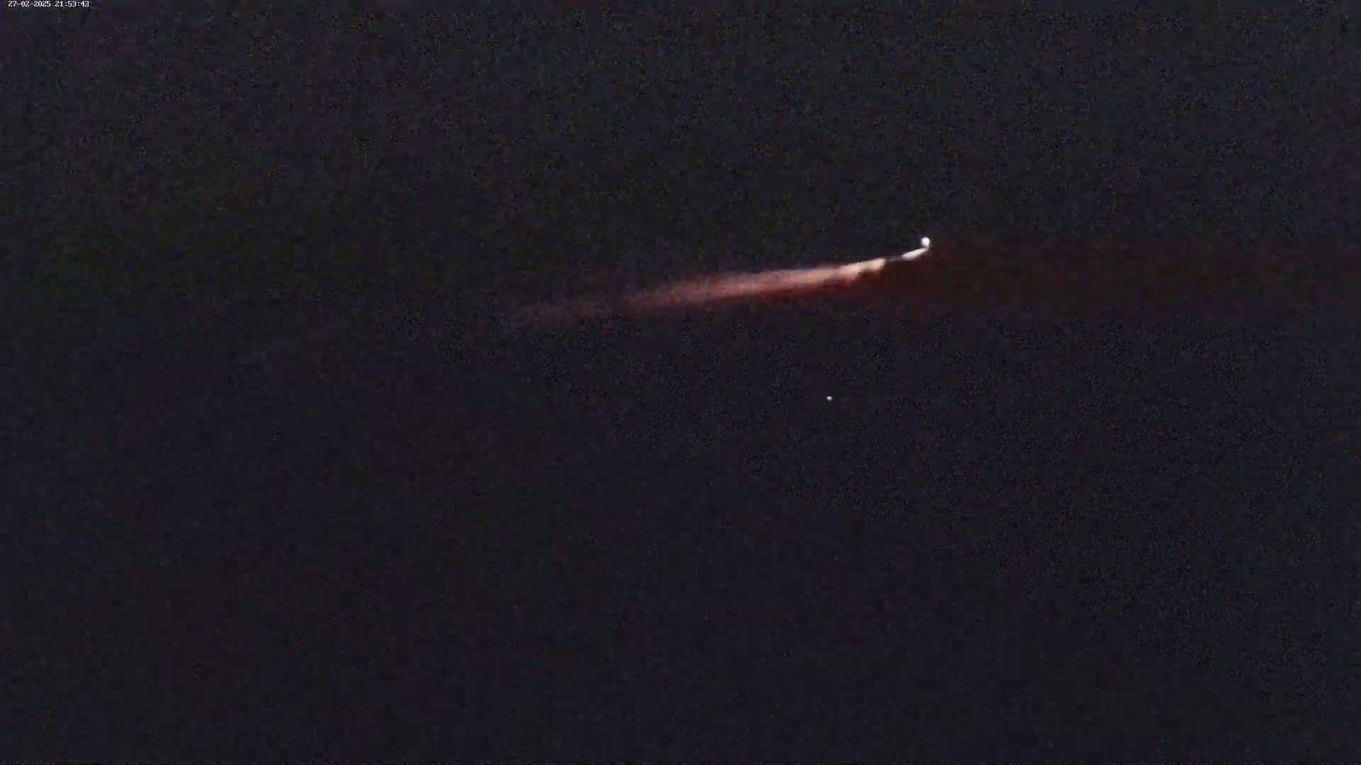 A long, reddish streak of light appears in a dark night sky, possibly a meteor or rocket trail.