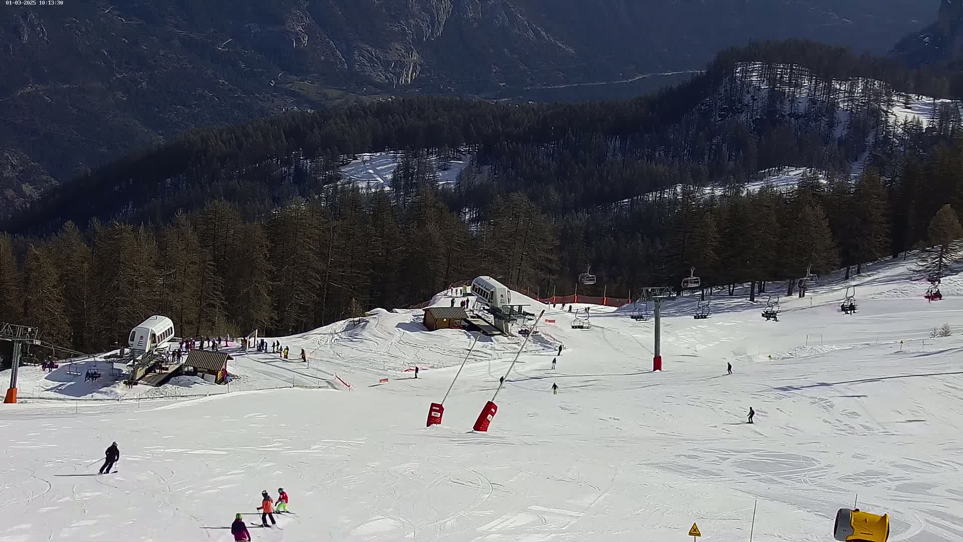 A sunny snow-covered ski slope with several skiers and snowboarders, ski lifts, and a mountainside forest in the background.