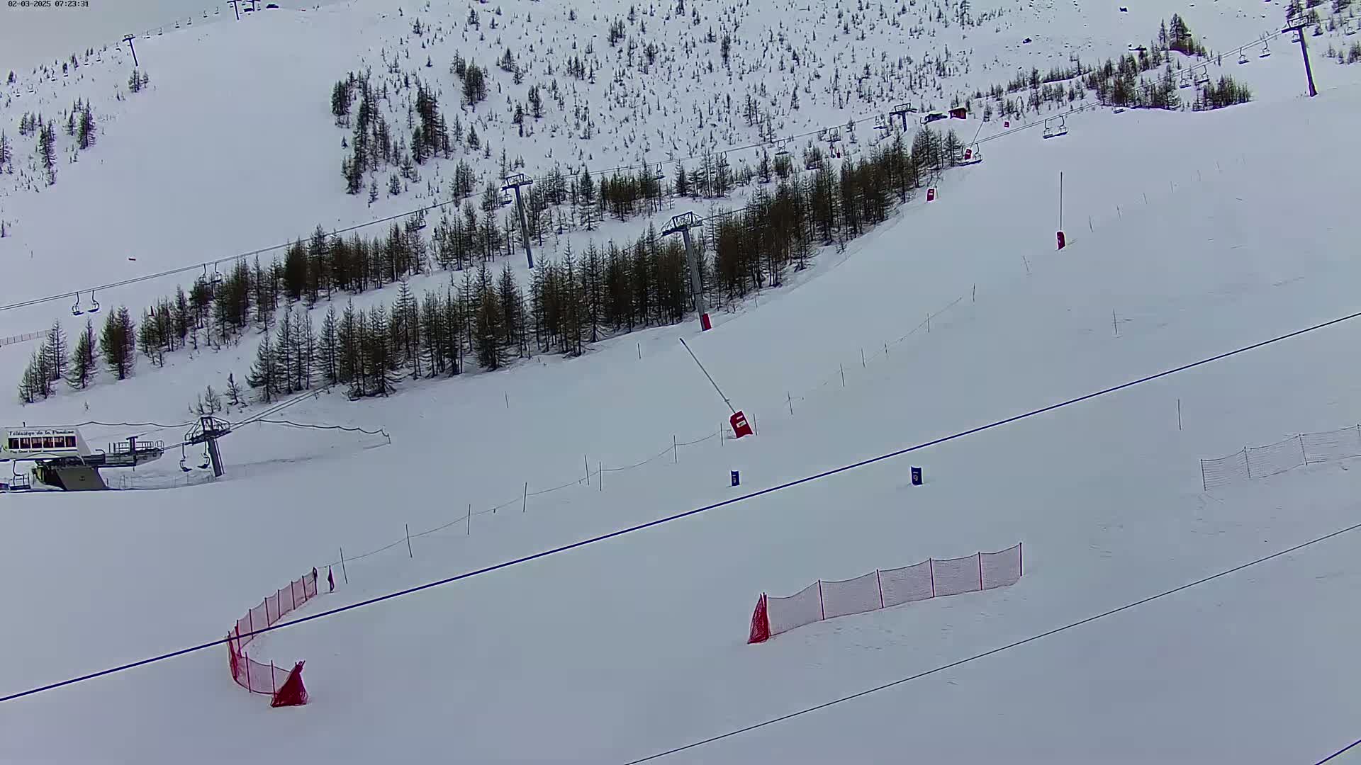 A snow-covered ski slope with several ski lifts, sparse evergreen trees, and red safety nets.