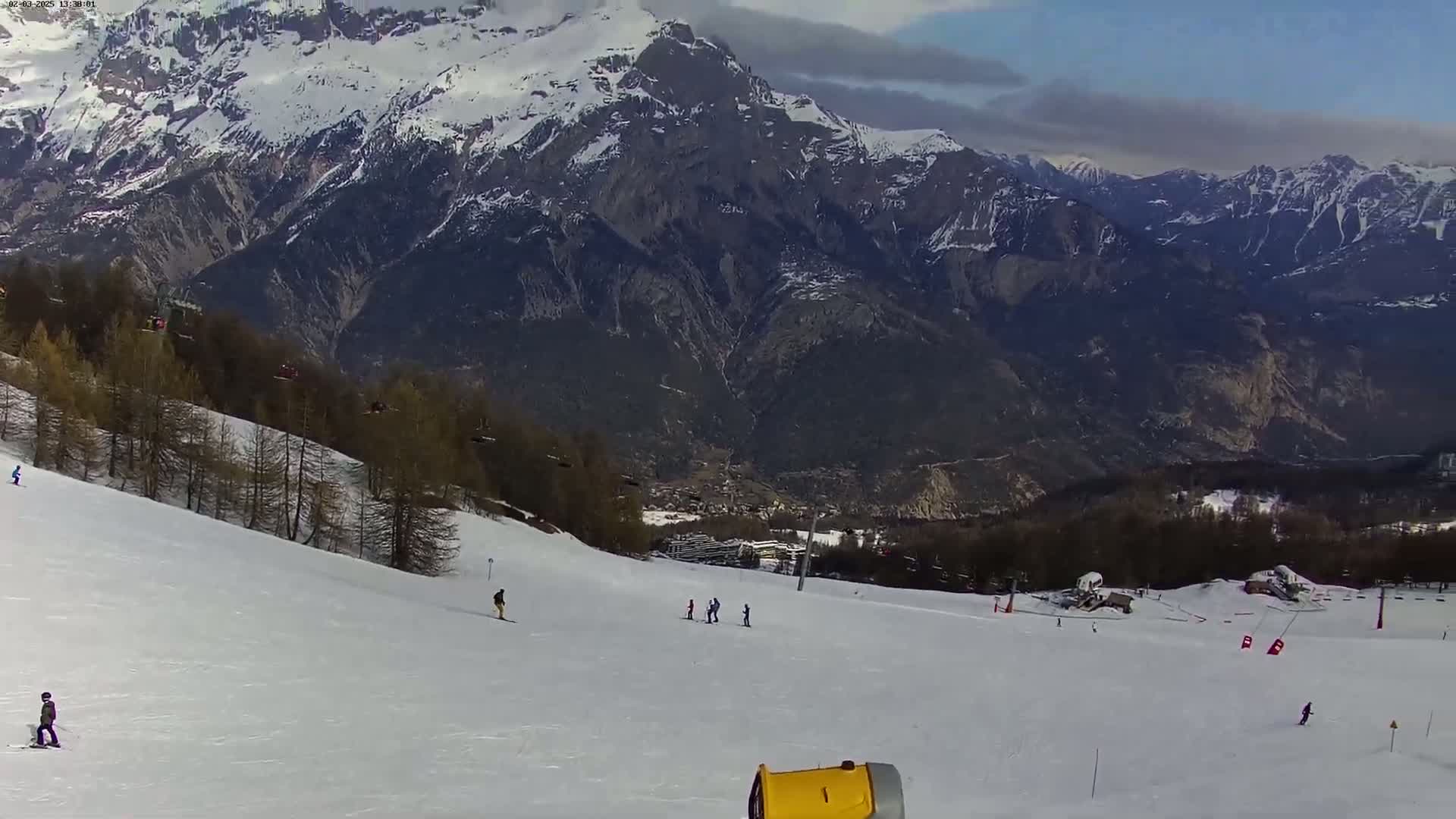 A snow-covered ski slope with several skiers descends toward a valley and a mountain range under a partly cloudy sky.
