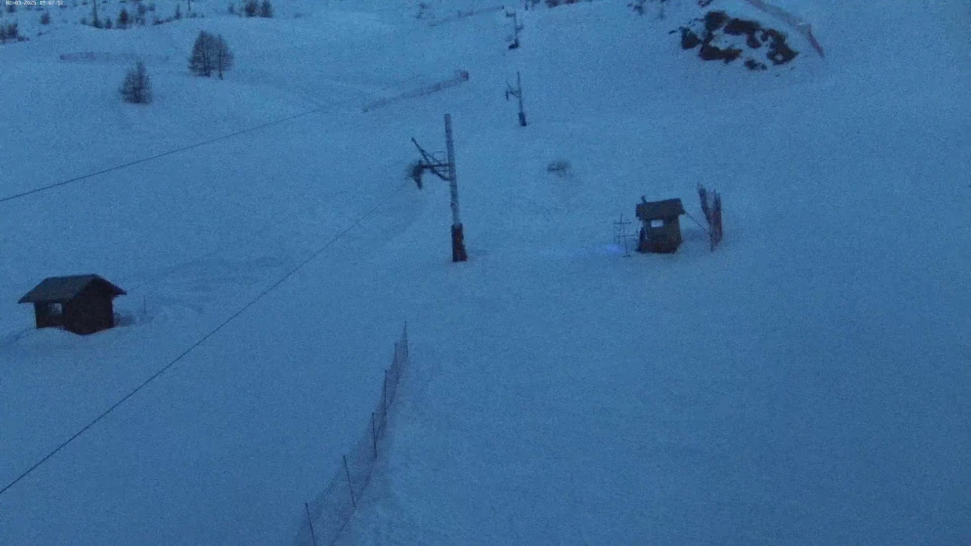 A snow-covered ski slope at twilight features two small wooden structures, a ski lift, and a section of fencing.