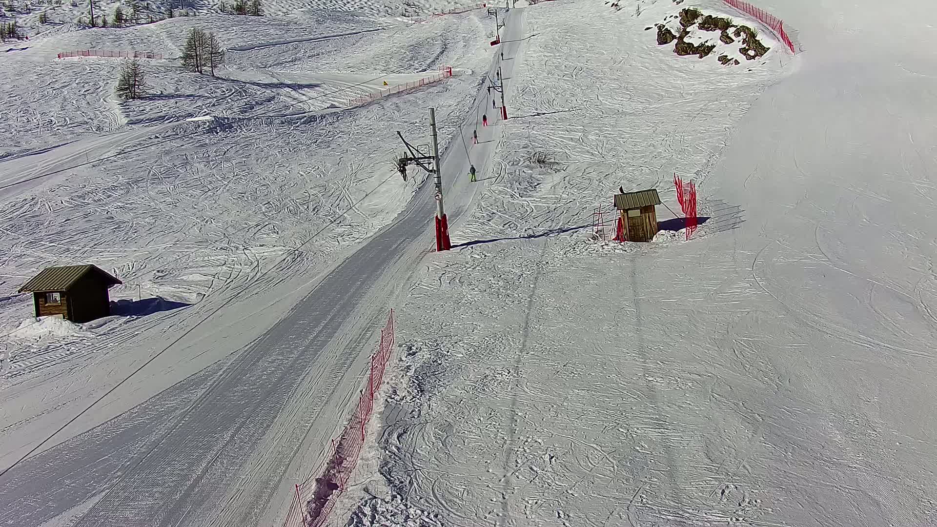 A ski slope with several skiers on a sunny day, featuring two small wooden structures and red fencing along the sides.
