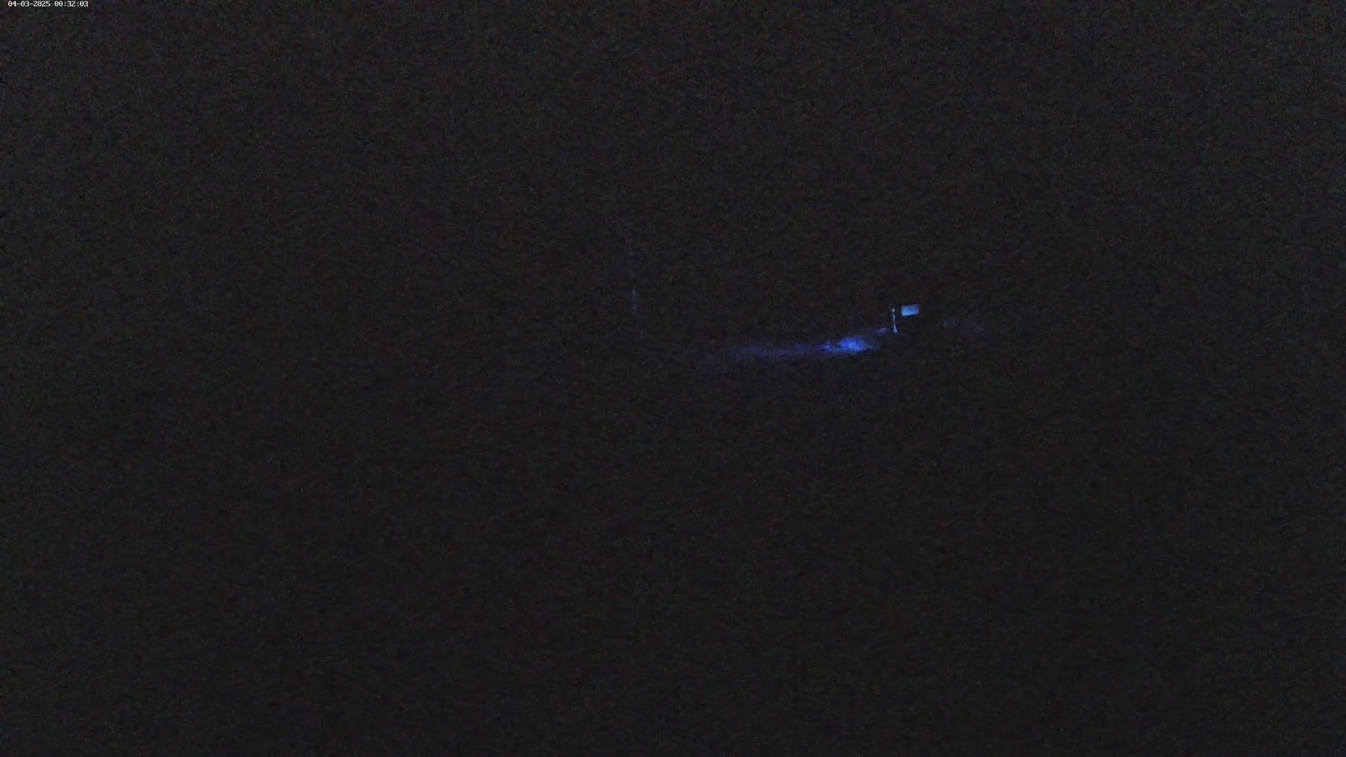 A dark, nighttime scene shows a faint blue light emanating from a distant object or structure.