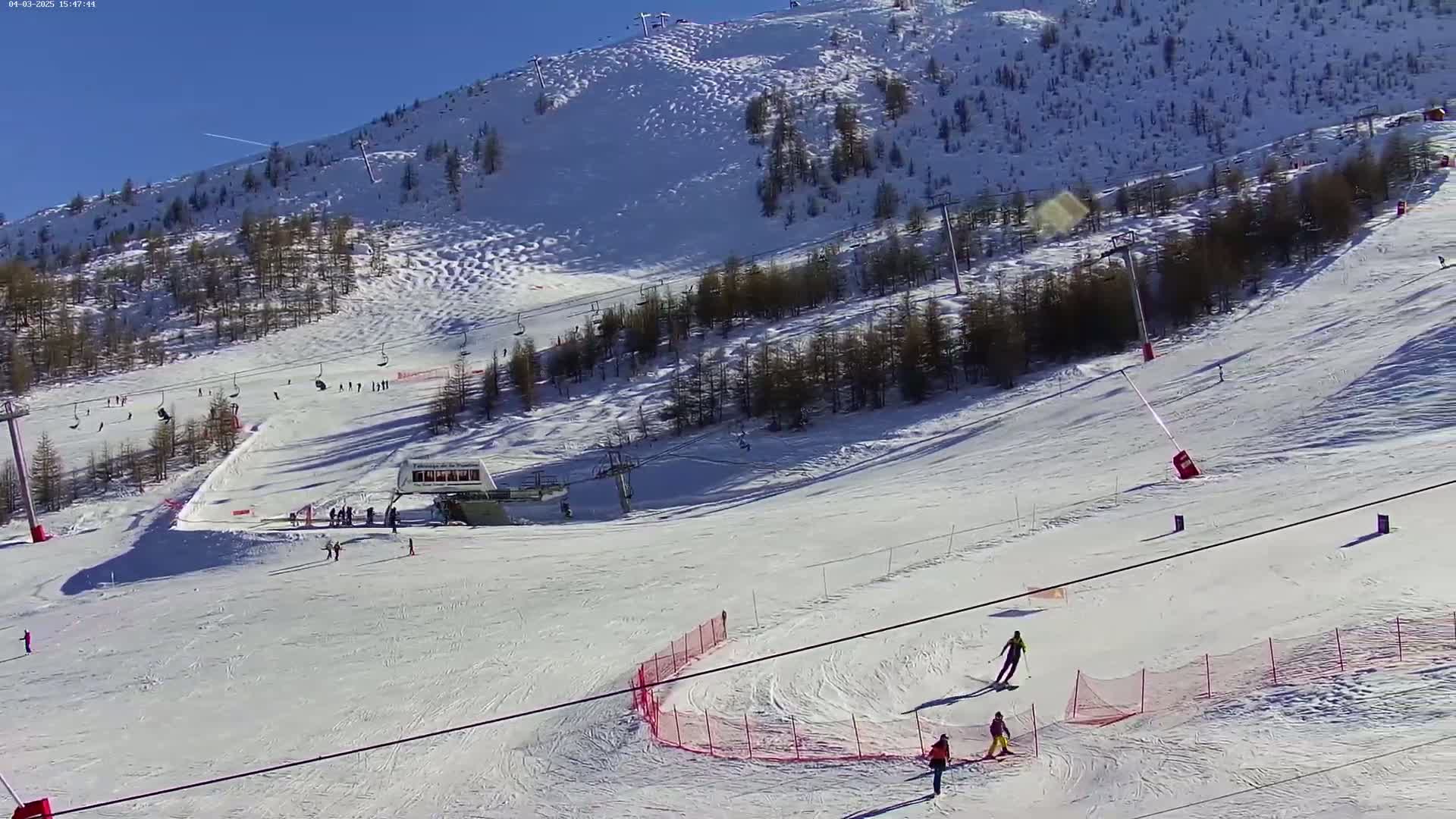 A sunny, snow-covered ski slope with several skiers and chairlifts, surrounded by evergreen trees on a mountainside.