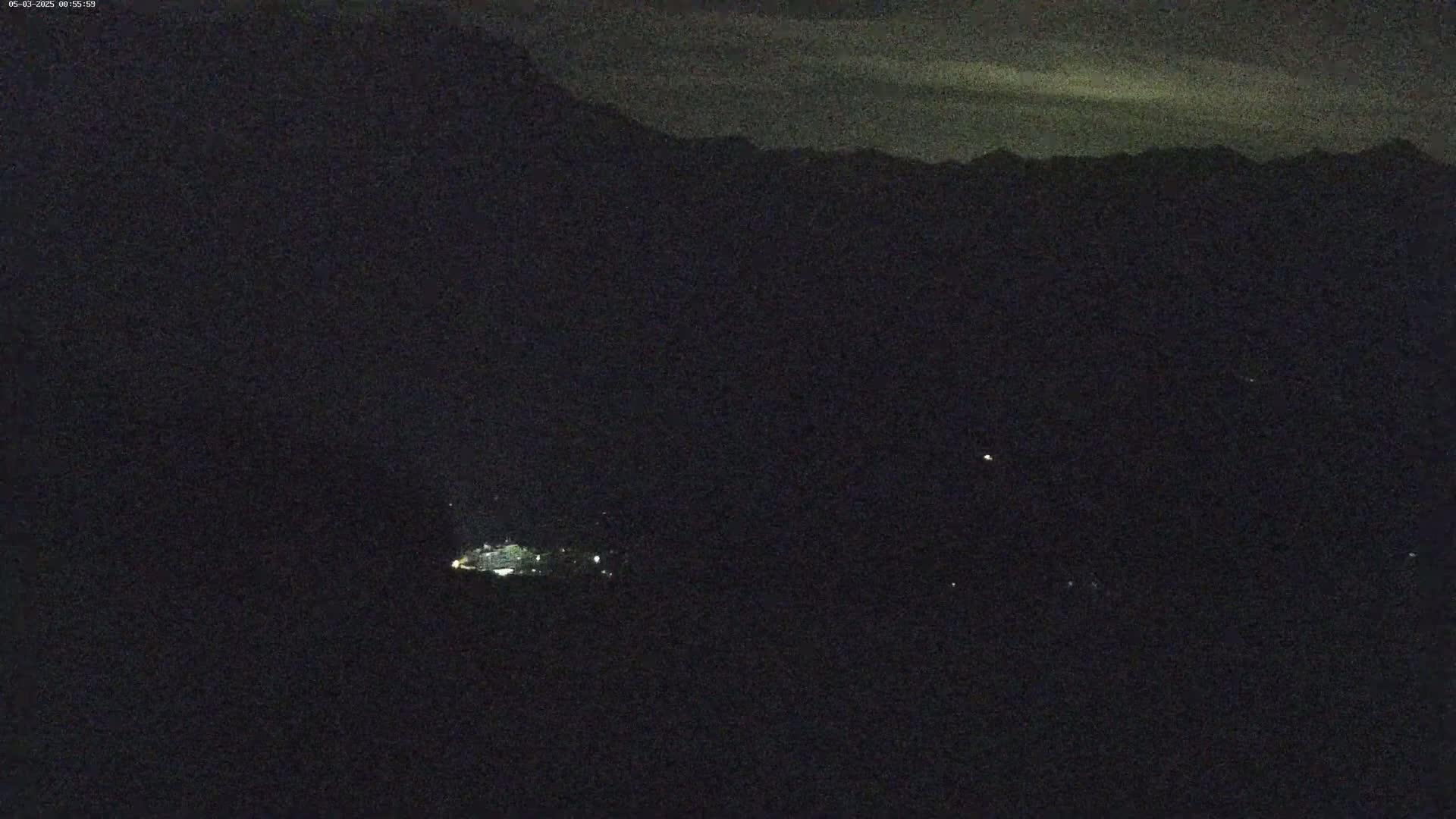 A nighttime long shot shows a dimly lit town nestled among dark mountains under a slightly cloudy sky.