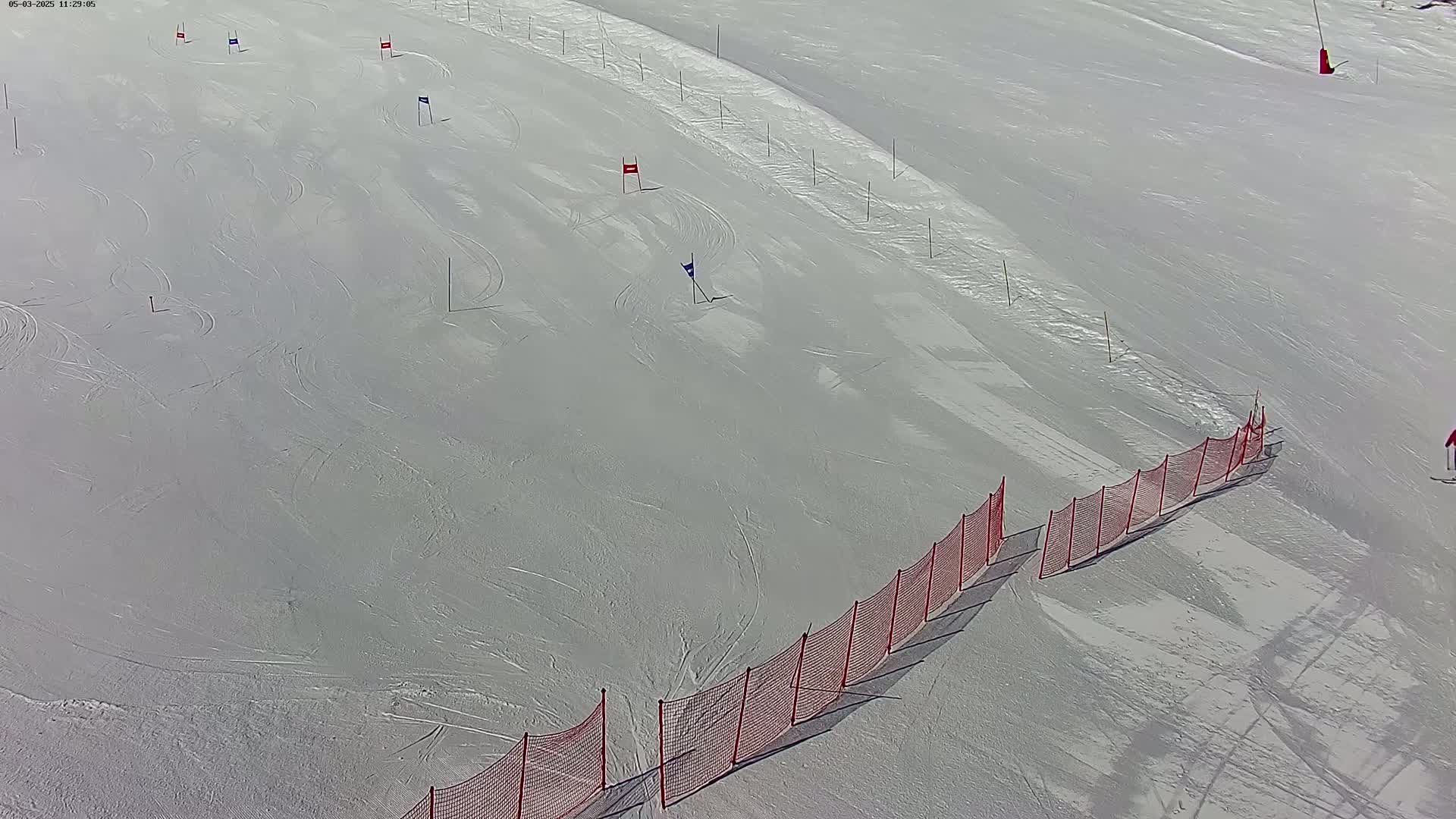 An aerial view shows a ski slope with a red safety net along one side, several flags marking a course, and a skier in the distance on a sunny day.