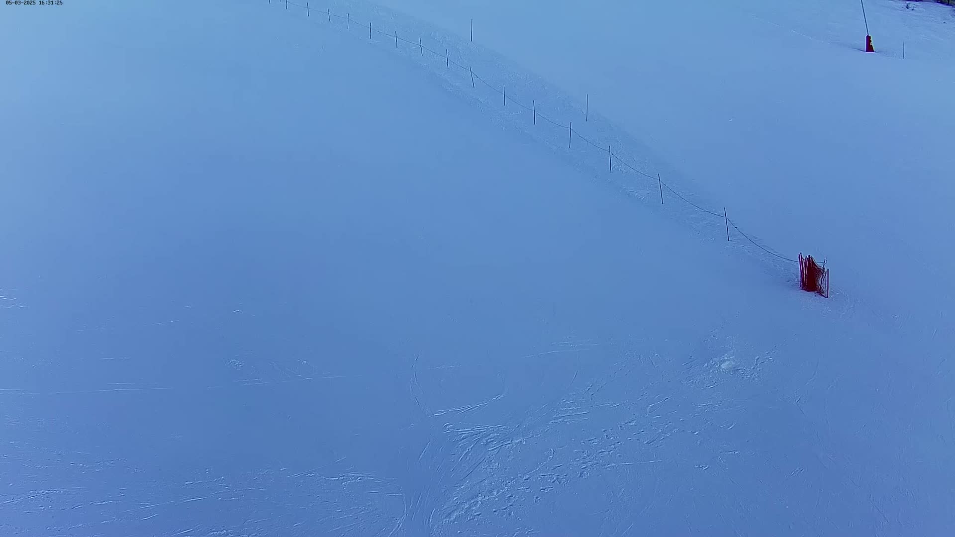 A snow-covered slope features a thin, meandering line of posts and wire, ending at a small, reddish-brown barrier.