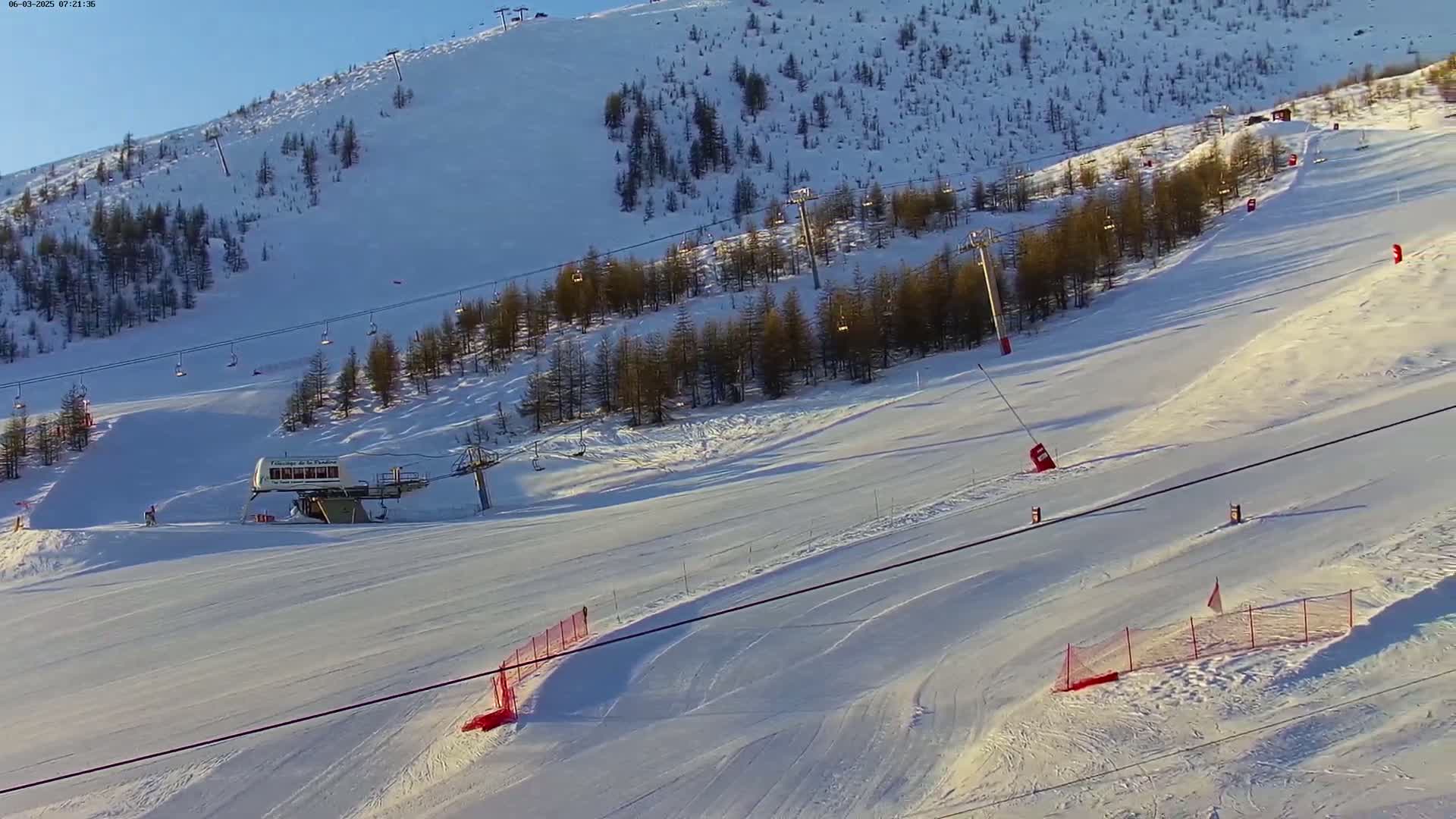 A sunny ski slope with a chairlift and several skiers is surrounded by snow-covered pine trees.