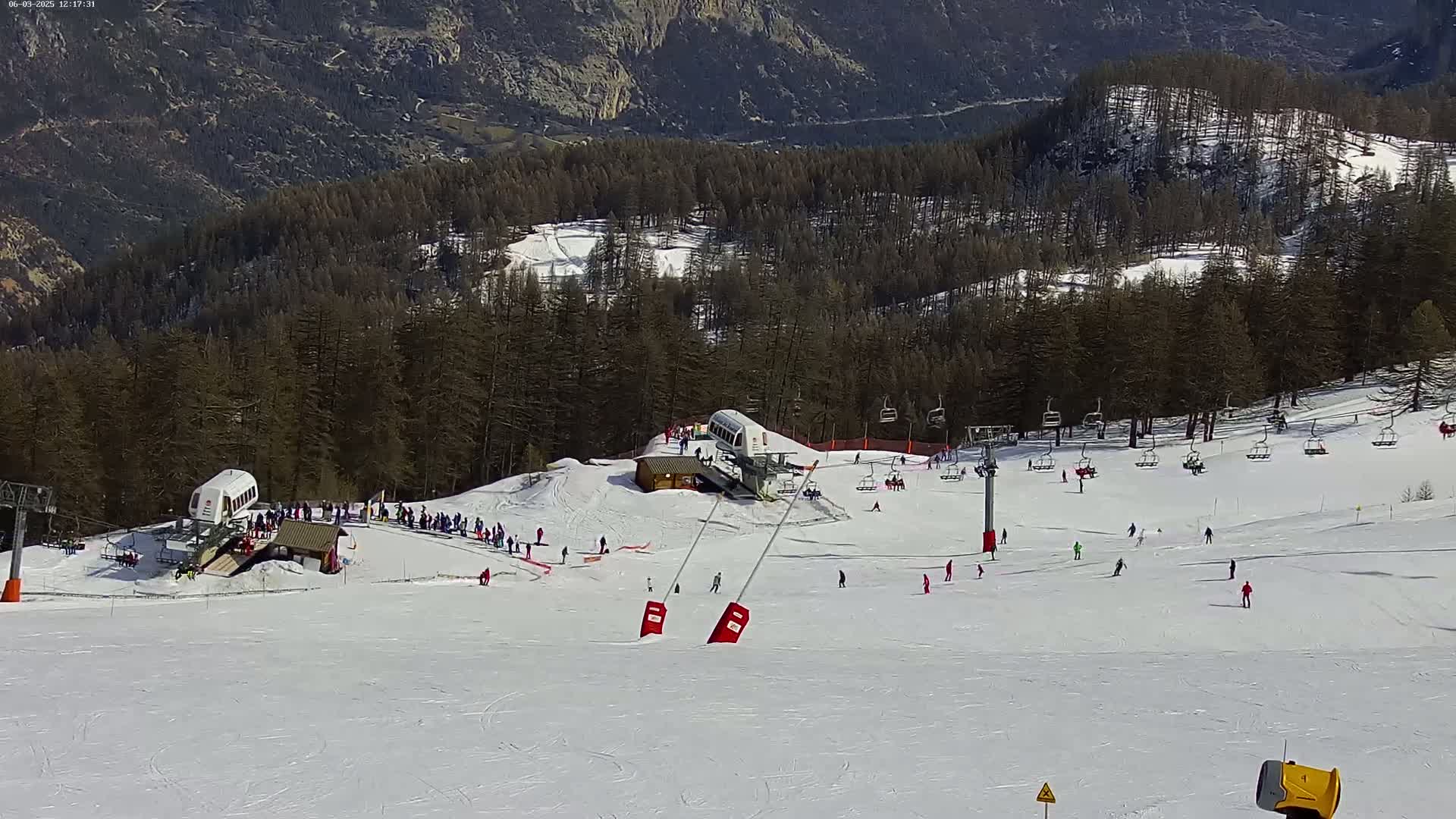 A sunny ski slope with several skiers and snowboarders, chairlifts, and a ski lift, is nestled in a mountainous forest.