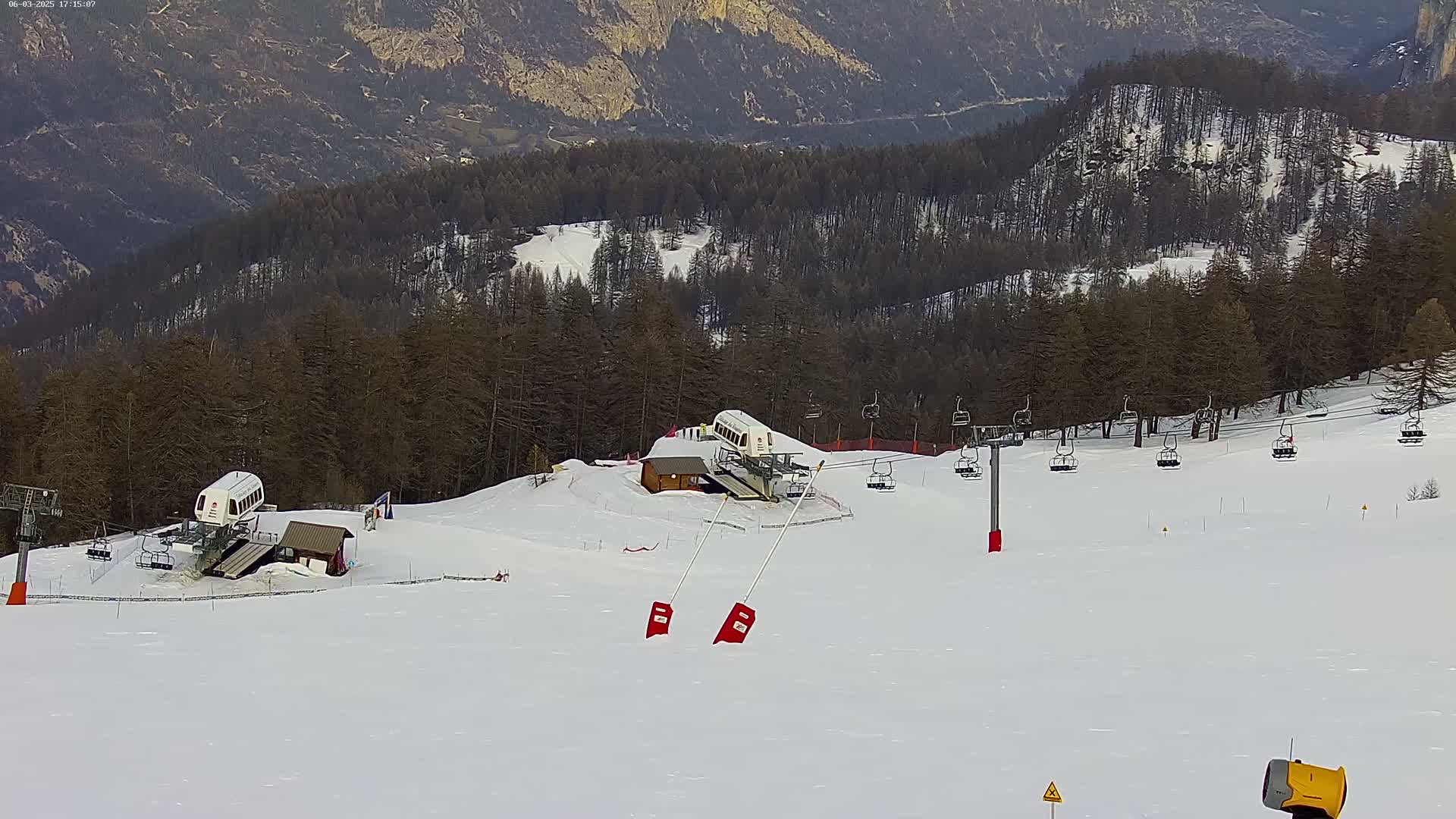 A snow-covered ski slope with two chairlifts, several empty chairlift cars, and a small building in the background, all set against a backdrop of a mountain range covered in dark evergreen trees.