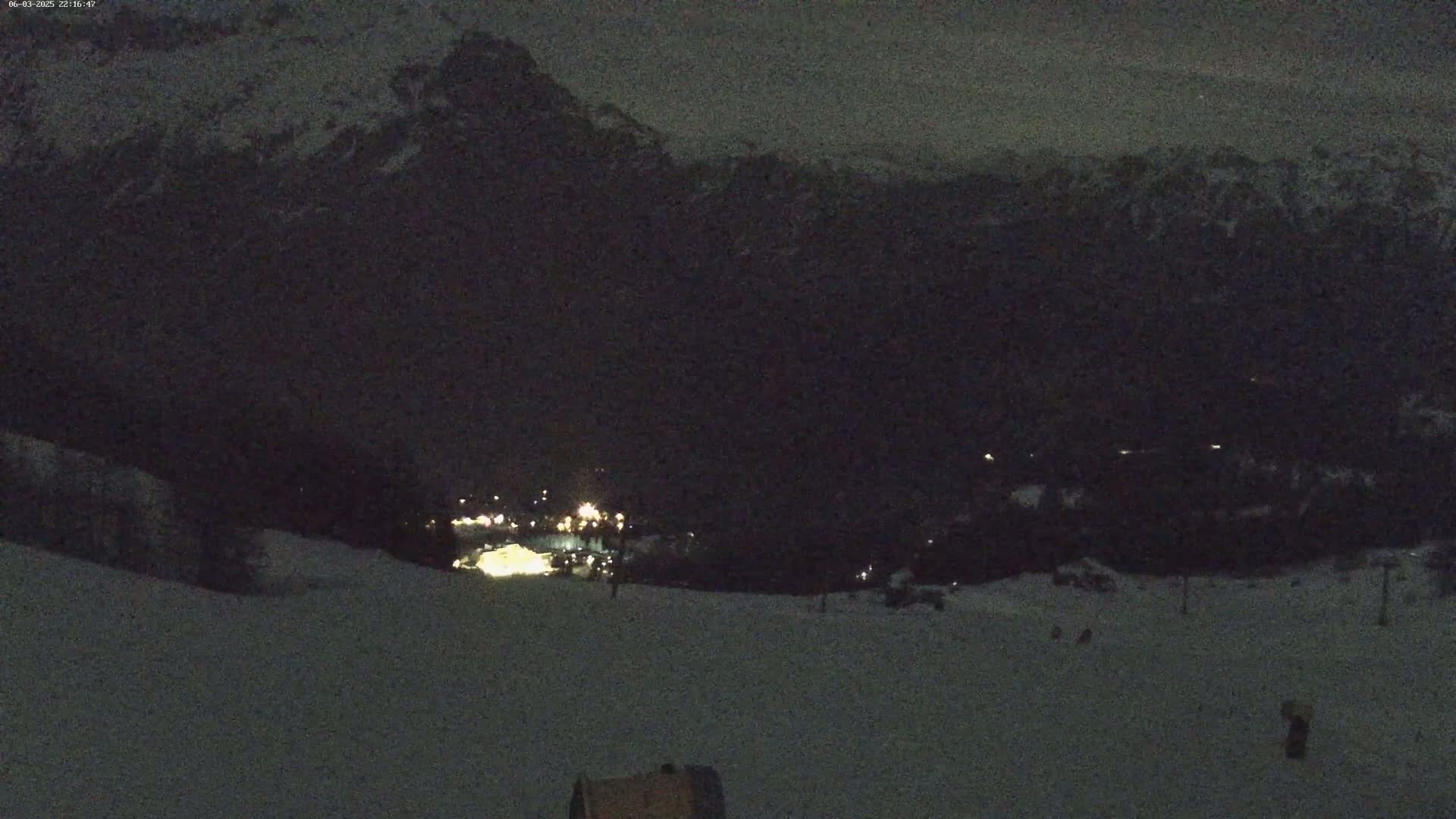 A nighttime view of a snow-covered slope leading down to a village illuminated by scattered lights, with dark mountains in the background under a dark sky.