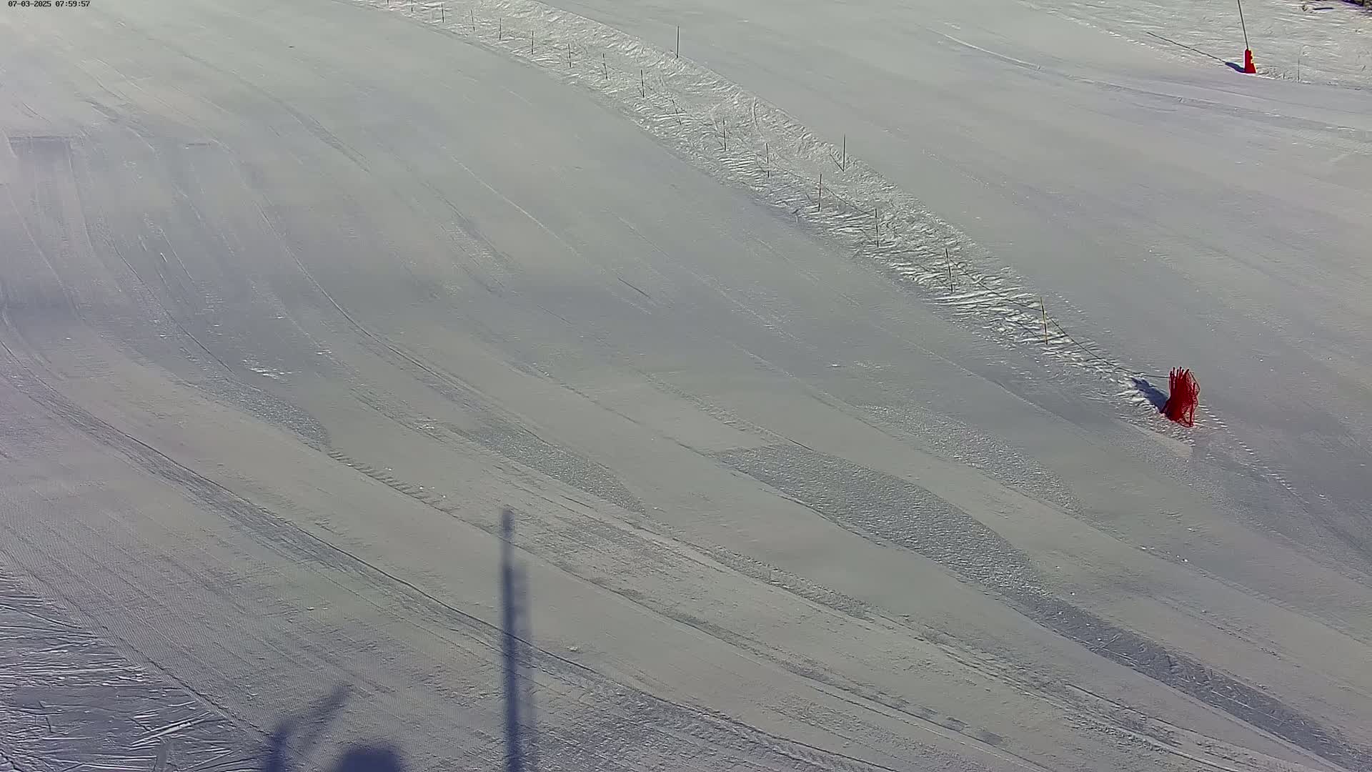 A snow-covered ski slope shows parallel tracks from skiers and snowboarders, with several orange markers and a long shadow cast on the snow under bright conditions.