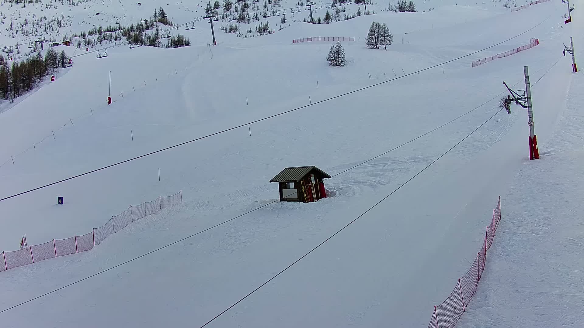 A snow-covered ski slope with a small wooden building, ski lift infrastructure, and red safety netting is visible under an overcast sky.
