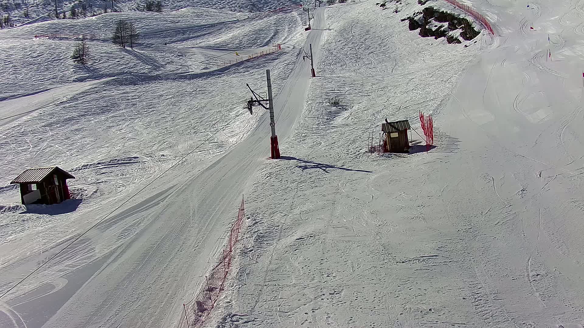A snow-covered ski slope with a chairlift, two small buildings, and red fencing on a sunny day.