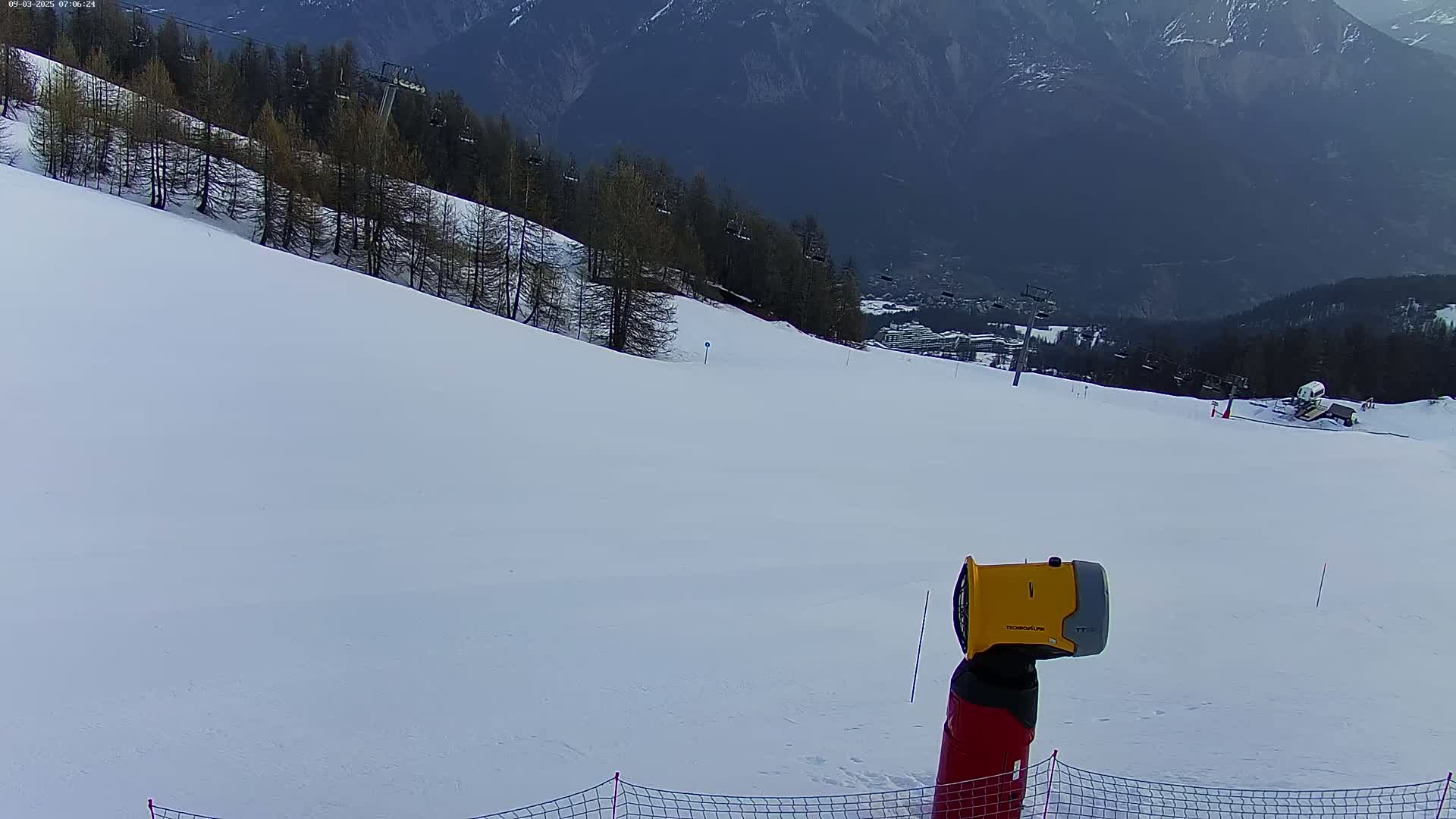 A snow-covered ski slope is seen from a low angle, with a snow gun in the foreground, a ski lift in the midground, and mountains in the background under a cloudy sky.