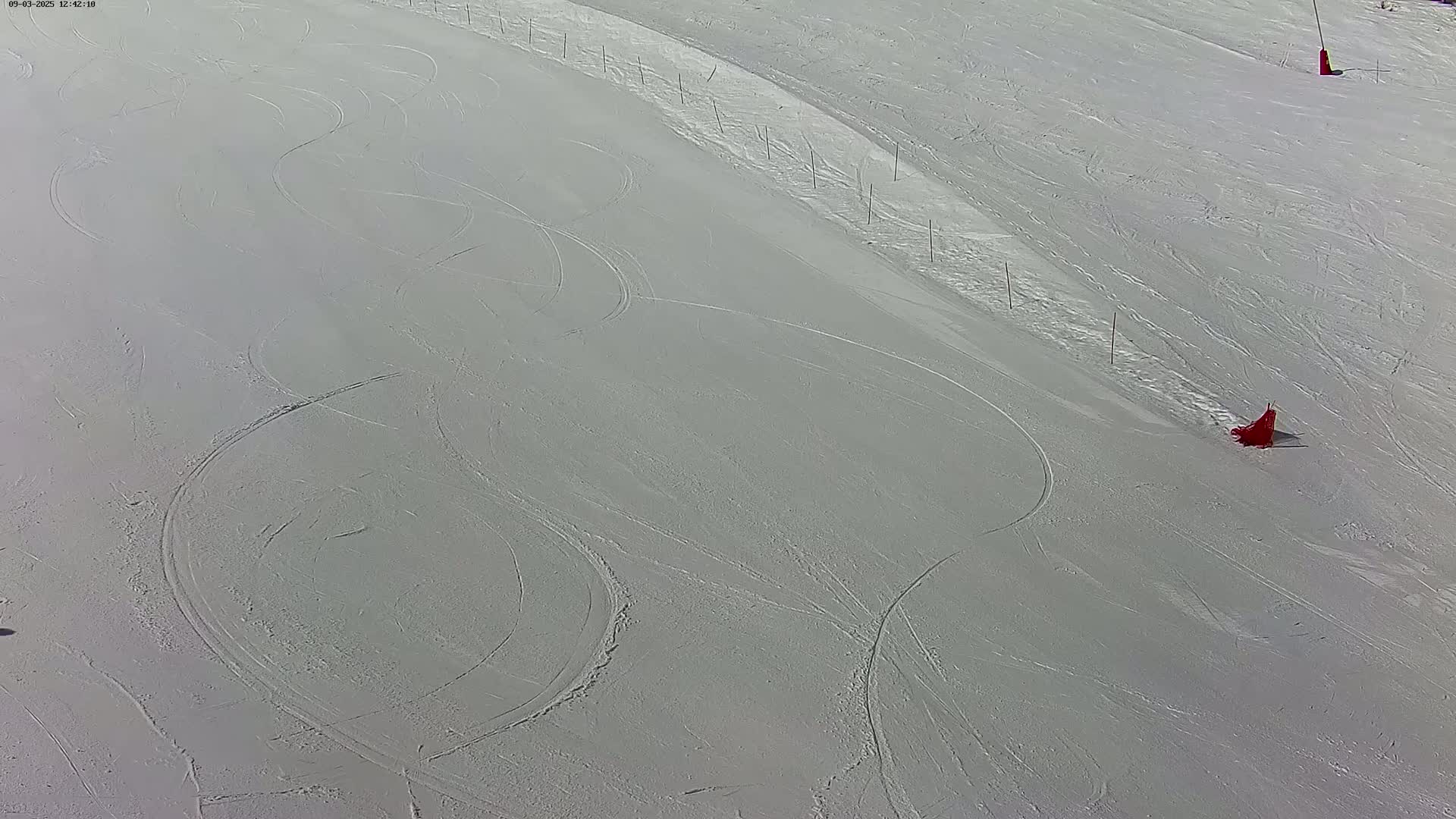 A snow-covered slope shows curving ski tracks and several red markers under a bright, sunny sky.