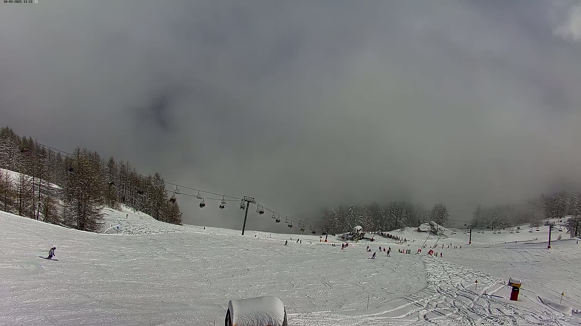 A snowy ski slope with a chairlift and several skiers and snowboarders is partially obscured by fog.