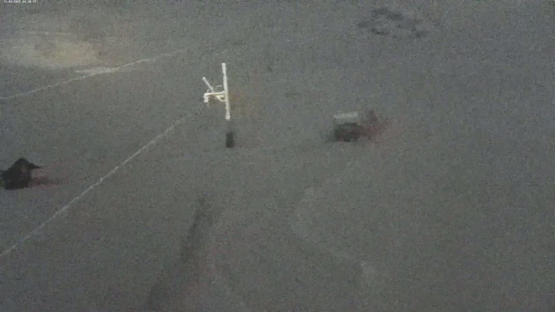 A blurry, dark gray asphalt surface shows a basketball hoop and indistinct shapes that may be vehicles or objects on the ground under an overcast sky.