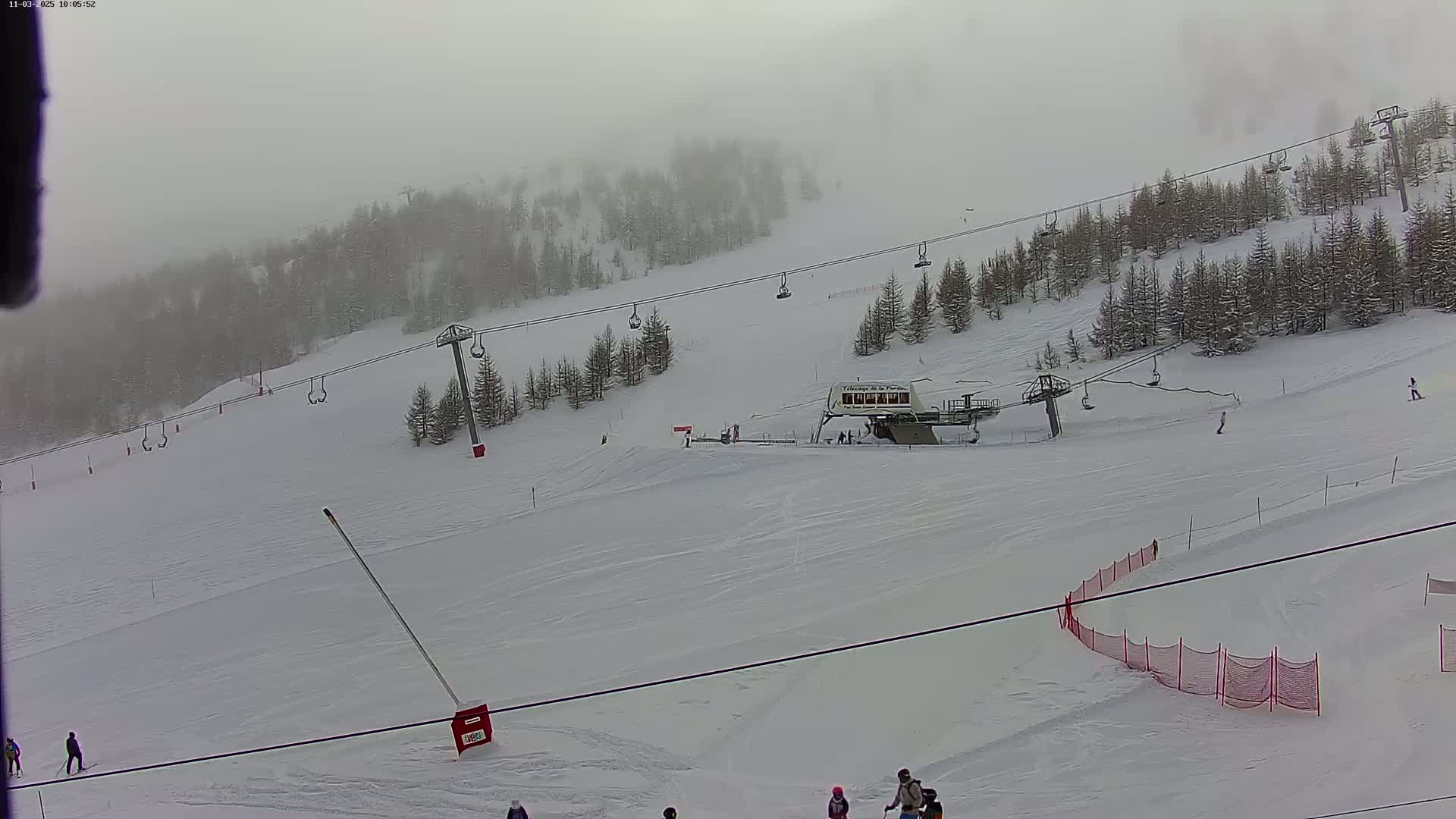 A snowy ski slope with chairlifts and a few skiers and snowboarders under foggy conditions.