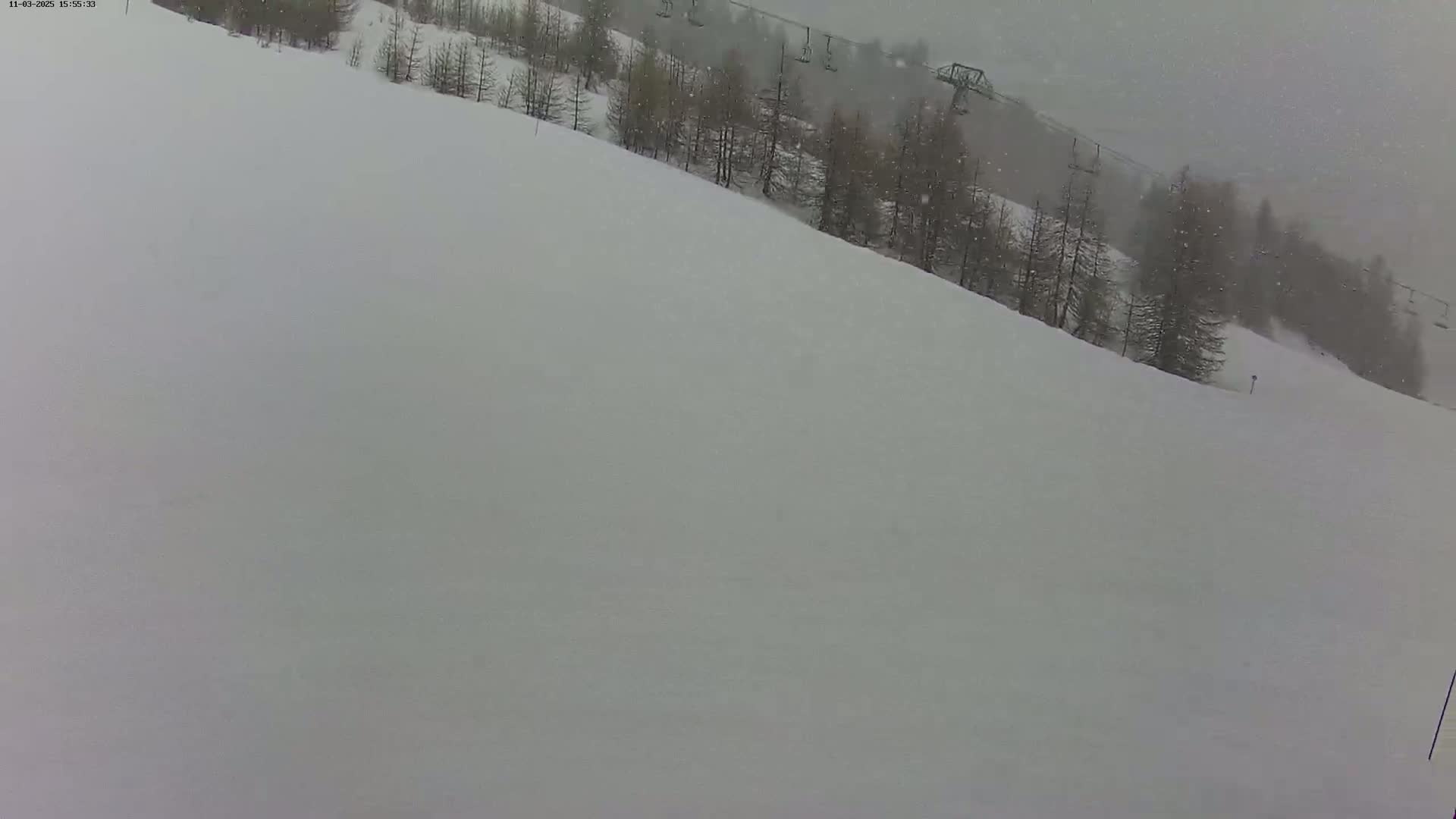 A snow-covered ski slope with sparsely wooded areas and chairlifts in the background, under snowy conditions.