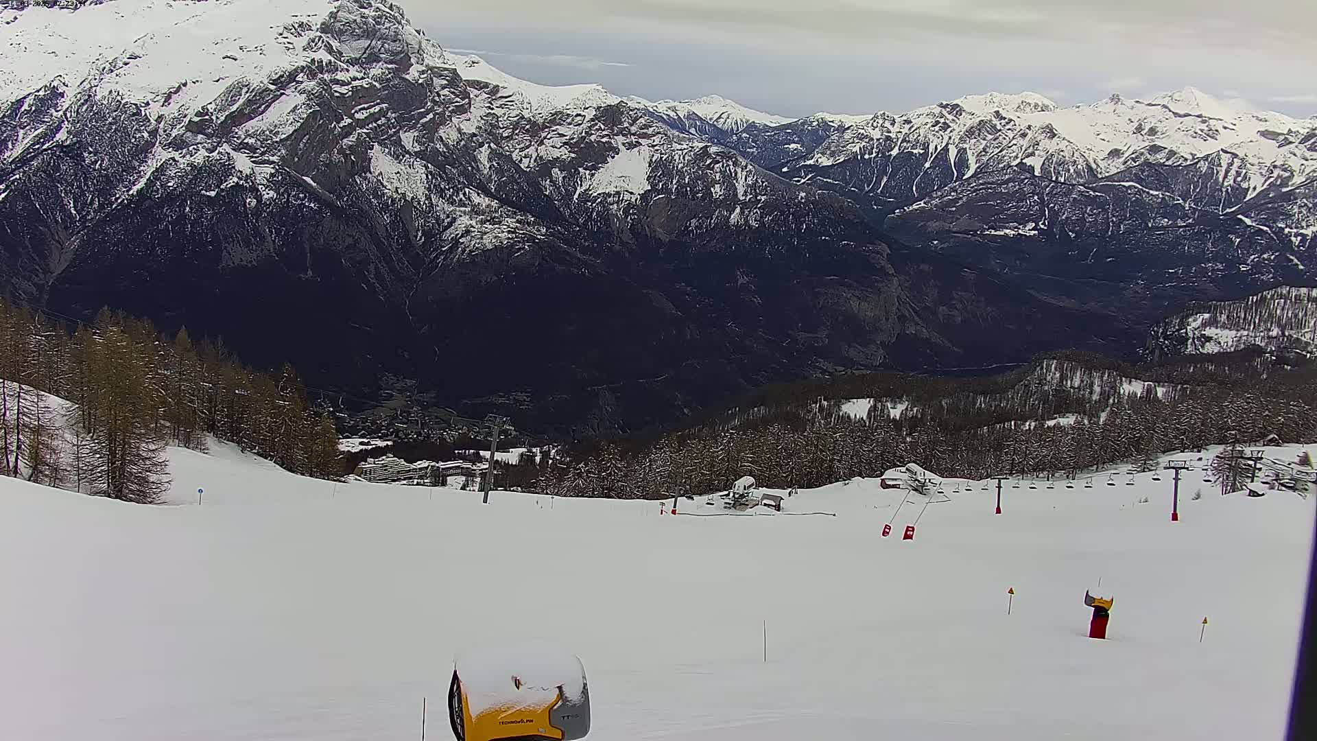 A snow-covered ski slope overlooks a valley with snow-capped mountains under a cloudy sky.