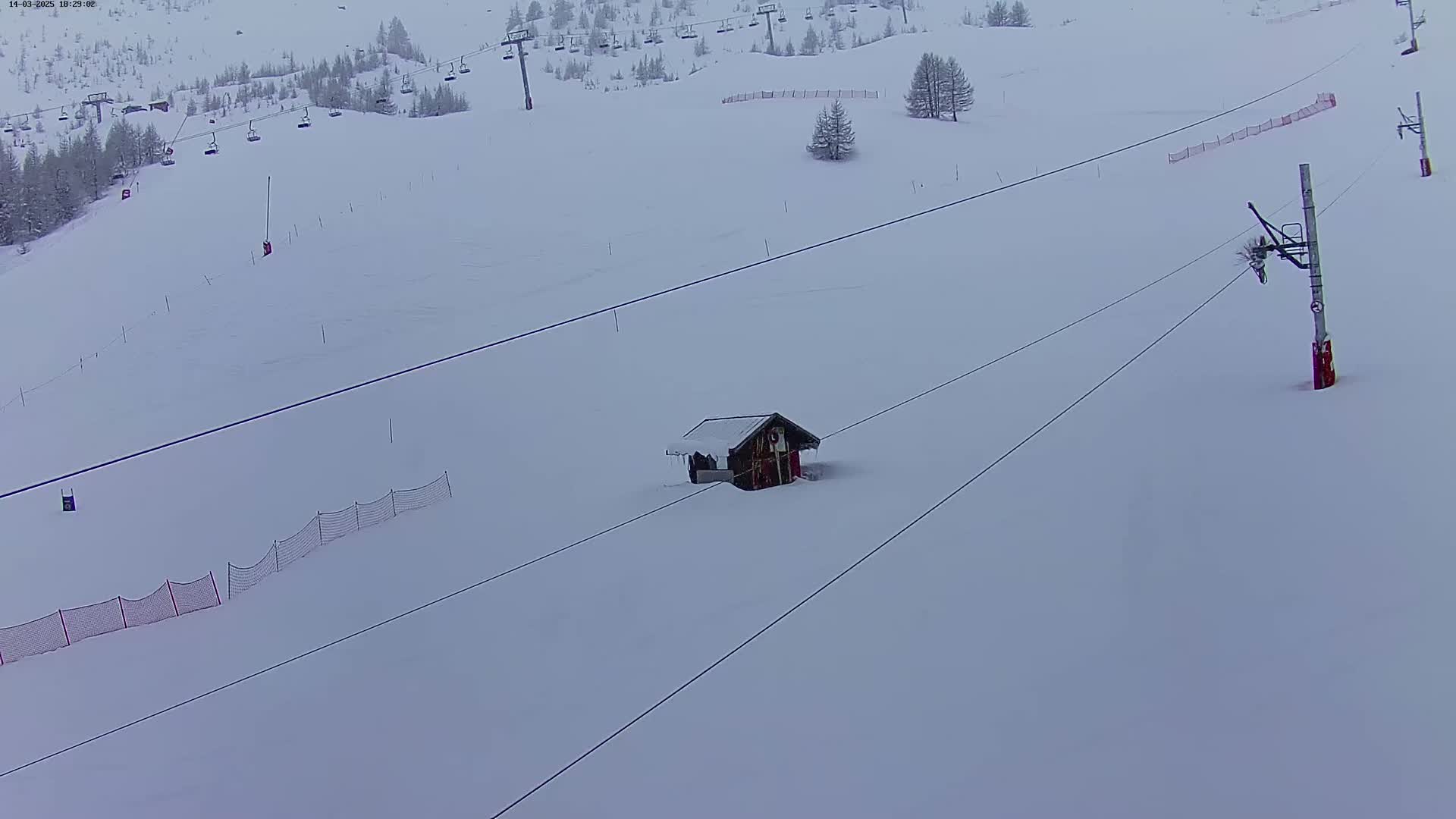 A snow-covered ski slope with a small cabin, ski lift infrastructure, and safety netting is visible under overcast skies.