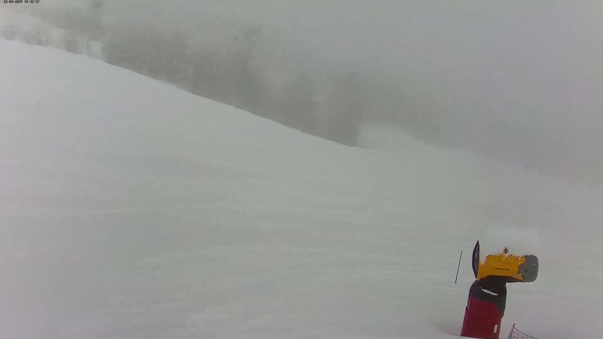 A snow-covered hill is visible through a heavy snowstorm, with a snow groomer partially buried in the snow in the foreground.