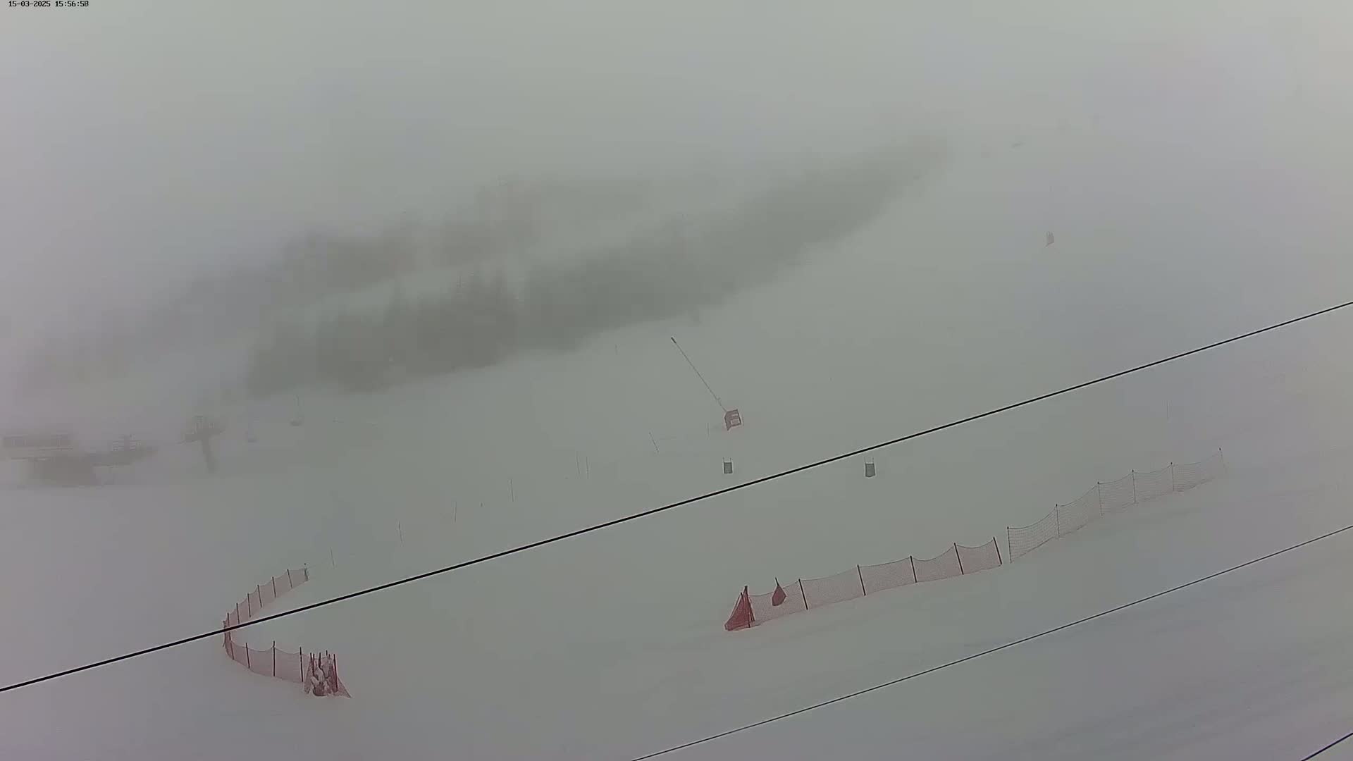 A snow-covered ski slope is mostly obscured by heavy fog, with some ski lift infrastructure and safety netting visible.