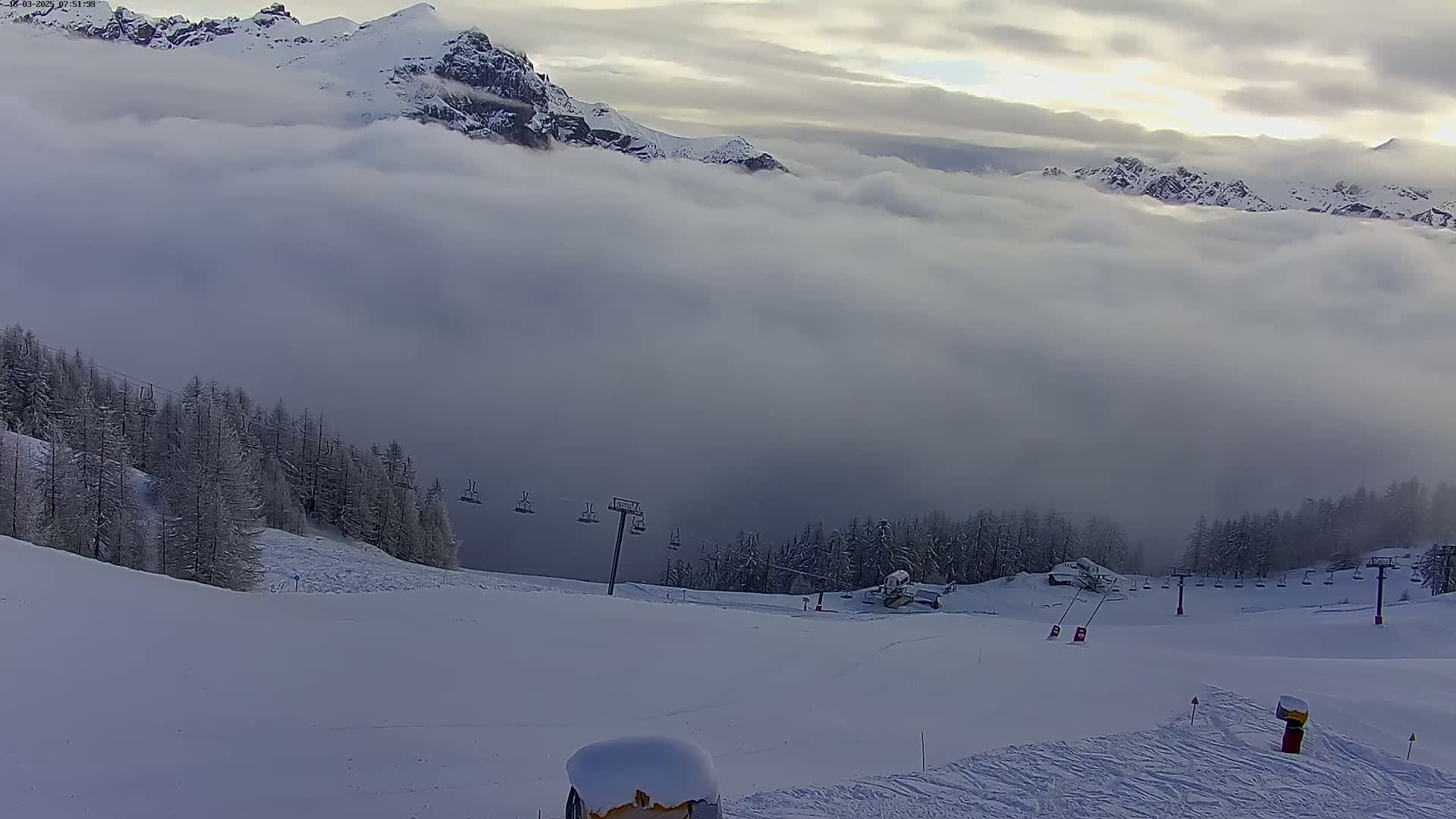 A snow-covered ski slope with a chairlift and snow-covered trees is partially obscured by a thick layer of fog or clouds, with snow-capped mountains visible in the background under an overcast sky.