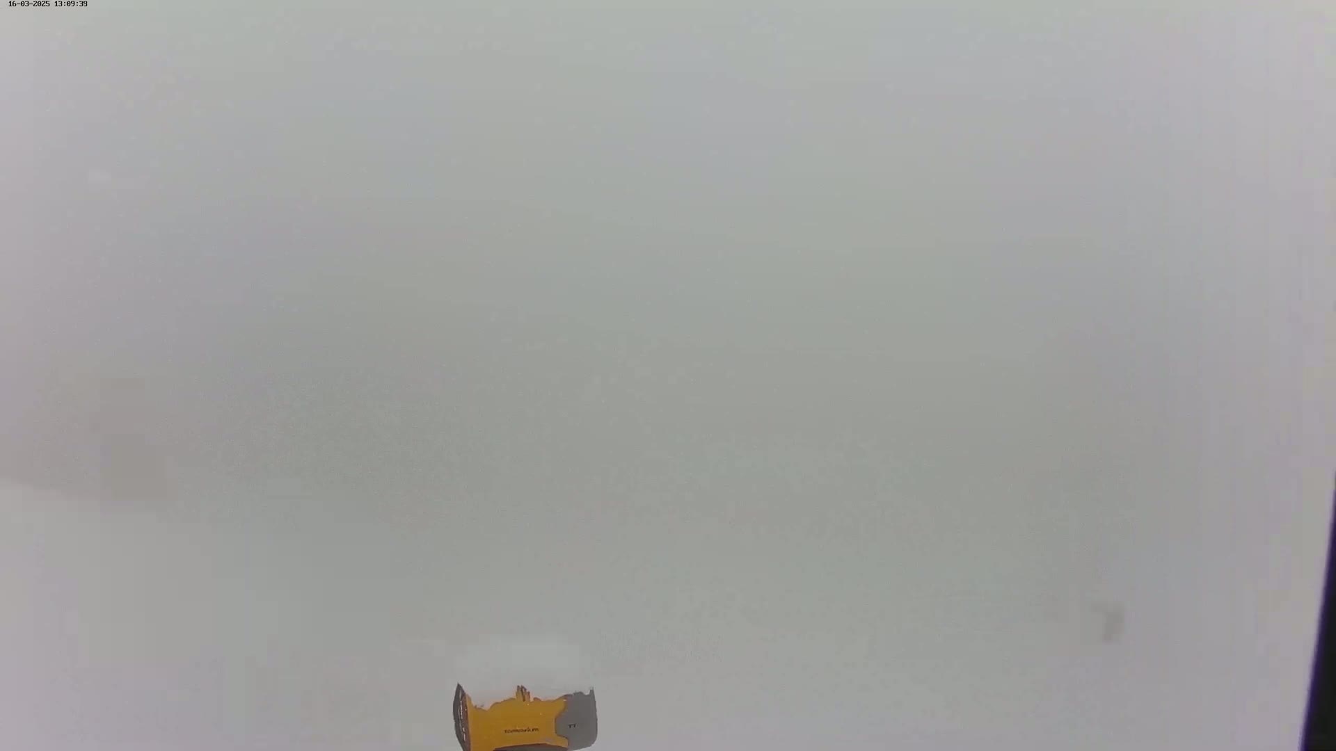 A small, yellow snowplow is visible in the lower center of a mostly obscured, snowy landscape under heavy overcast conditions.