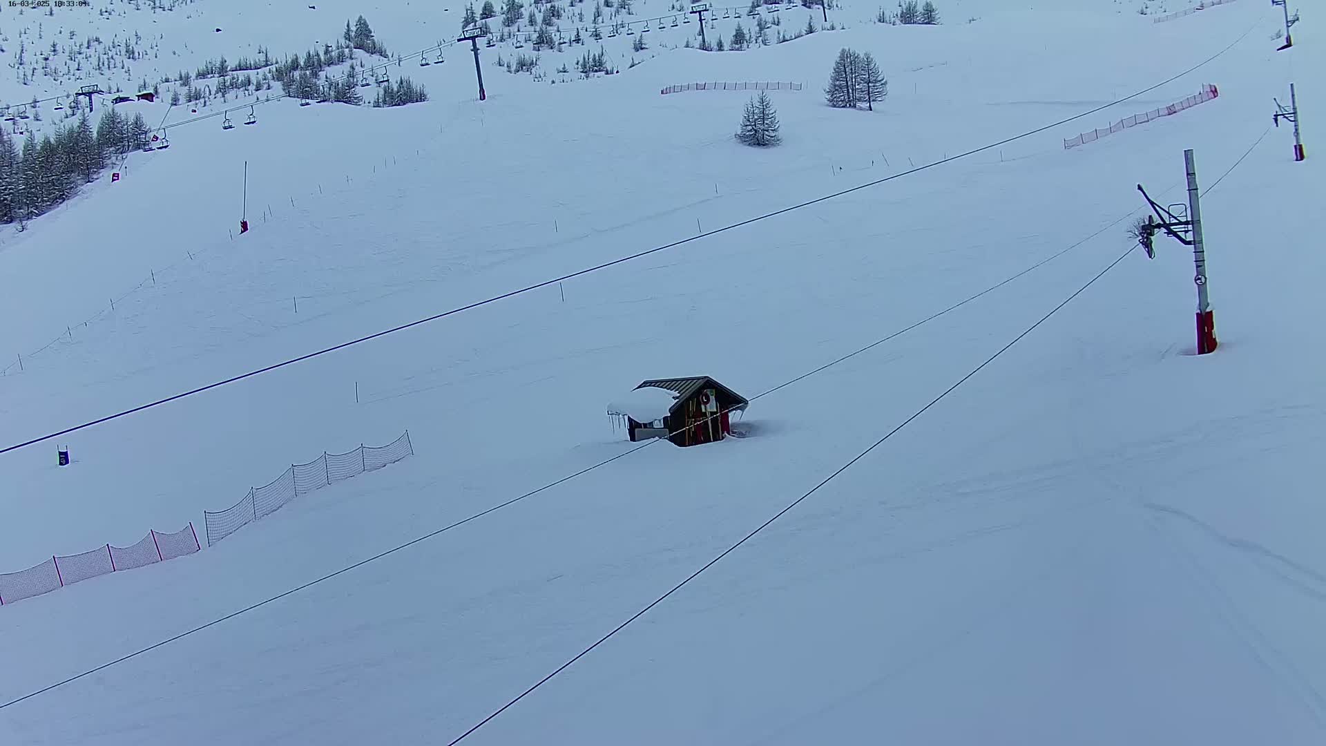 A snow-covered ski slope with a small building, ski lifts, and safety netting under an overcast sky.