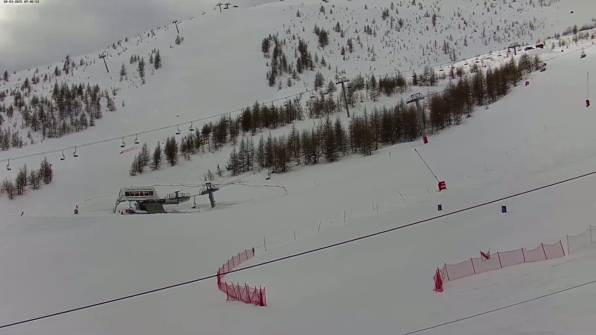 A snowy ski slope with several chairlifts, sparse evergreen trees, and red safety netting is visible under an overcast sky.