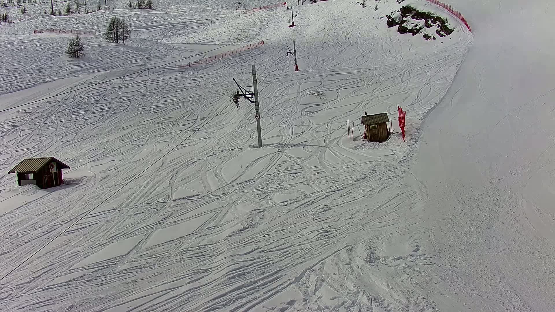 A snow-covered ski slope with two small buildings, ski lift supports, and numerous ski tracks under bright, sunny conditions.