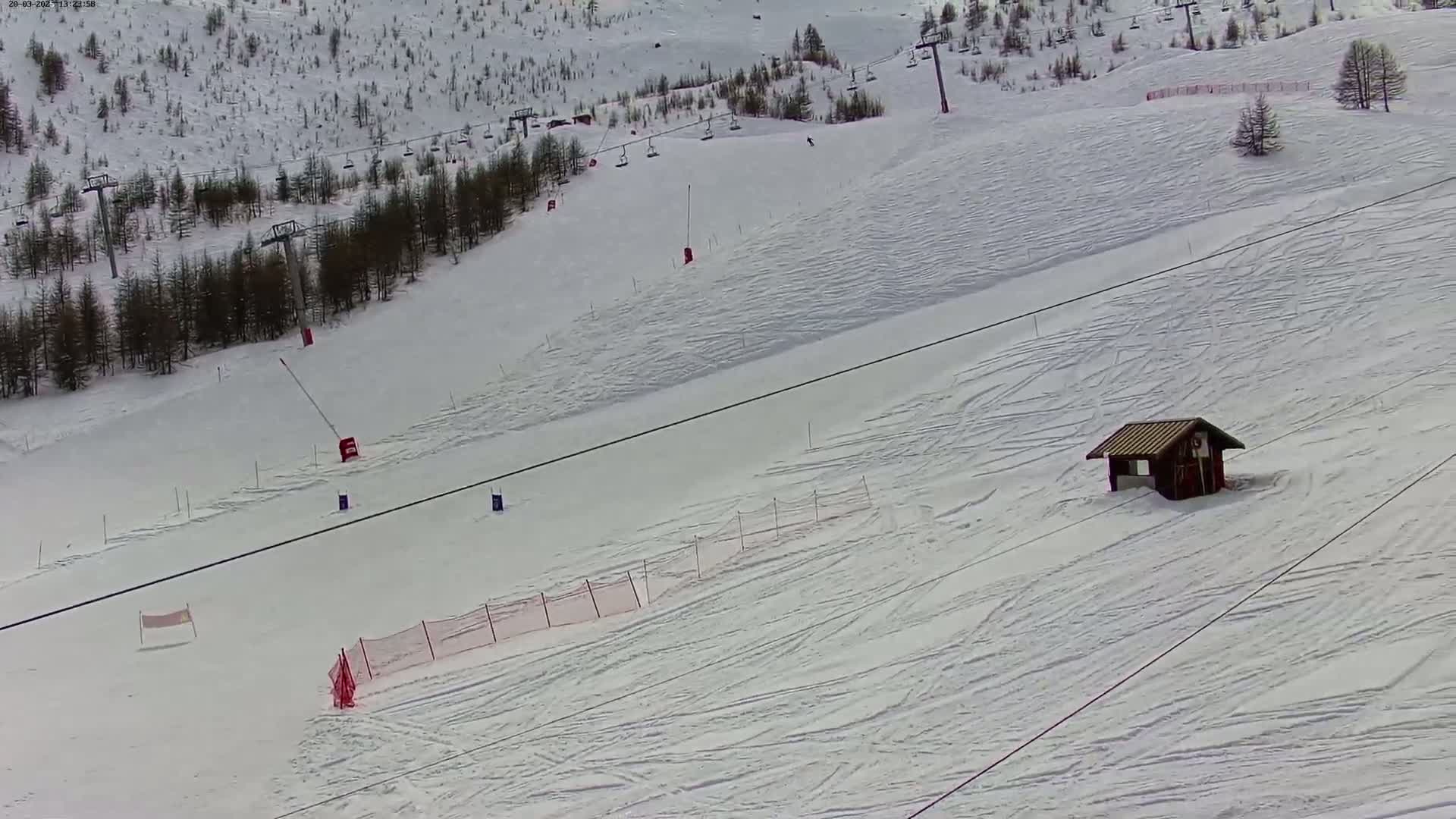 A snowy ski slope with several ski lifts, a small wooden building, and some safety netting is visible on an overcast day.