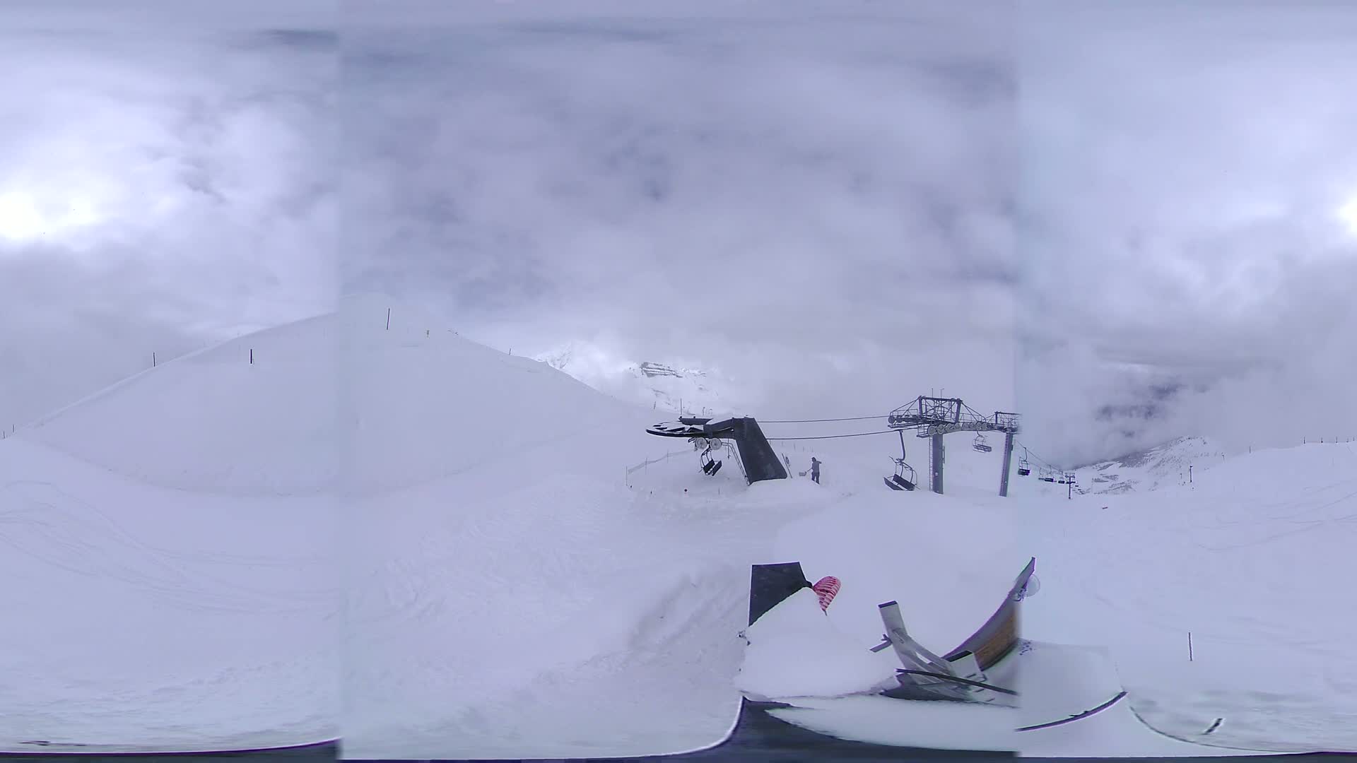 A snowy mountain landscape under cloudy skies shows a ski lift and a snow-covered terrain feature.