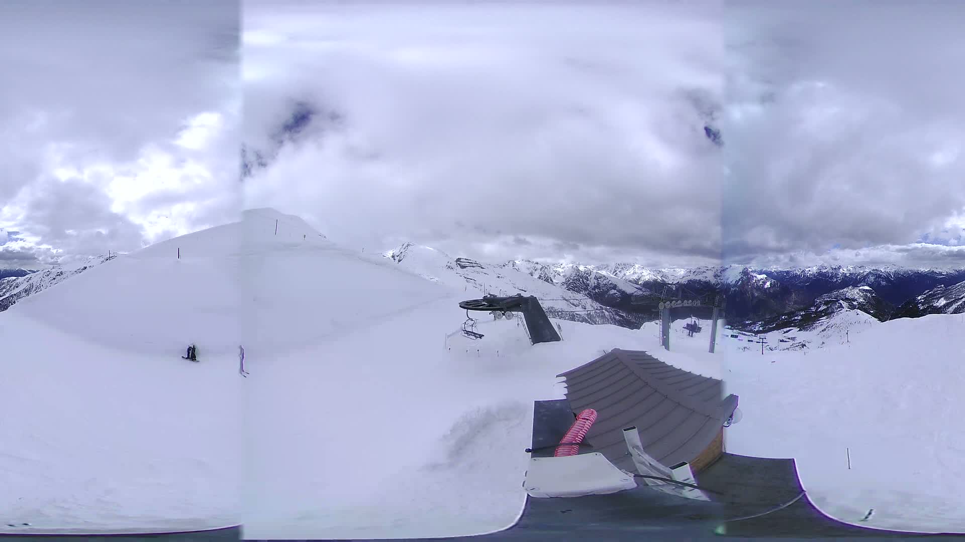 A panoramic view from a snowy mountaintop shows a ski lift, a small building, and several skiers on a cloudy day.