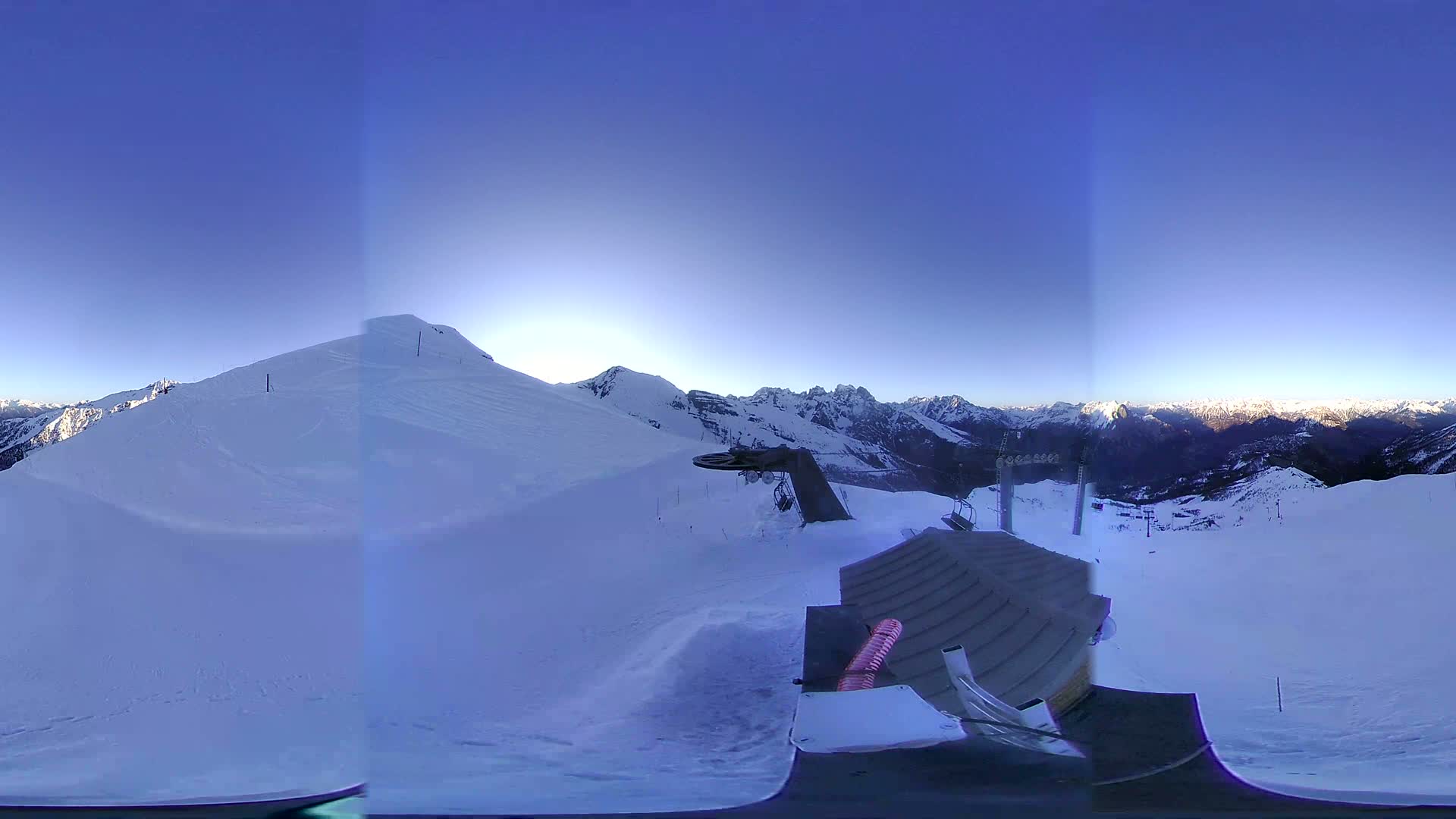 A snow-covered mountaintop with a clear blue sky, showing a ski lift and a panoramic view of other snow-capped mountains in the distance.
