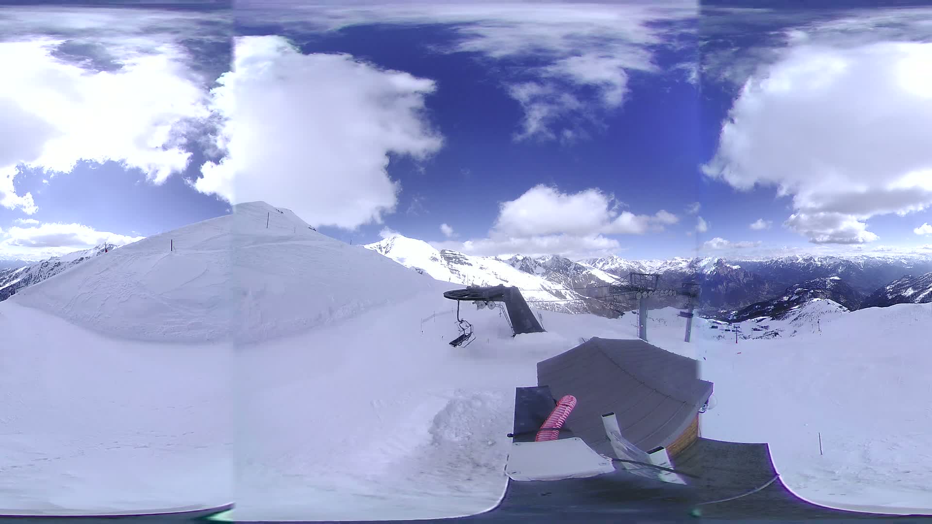 A panoramic view shows a snow-covered mountaintop with a ski lift and a partly cloudy blue sky.