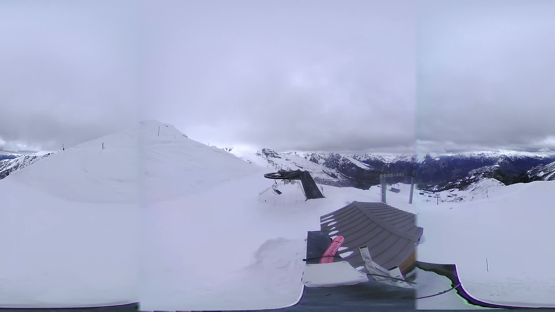 A snow-covered mountaintop with a ski lift and overcast skies is visible from a vantage point near a small building.