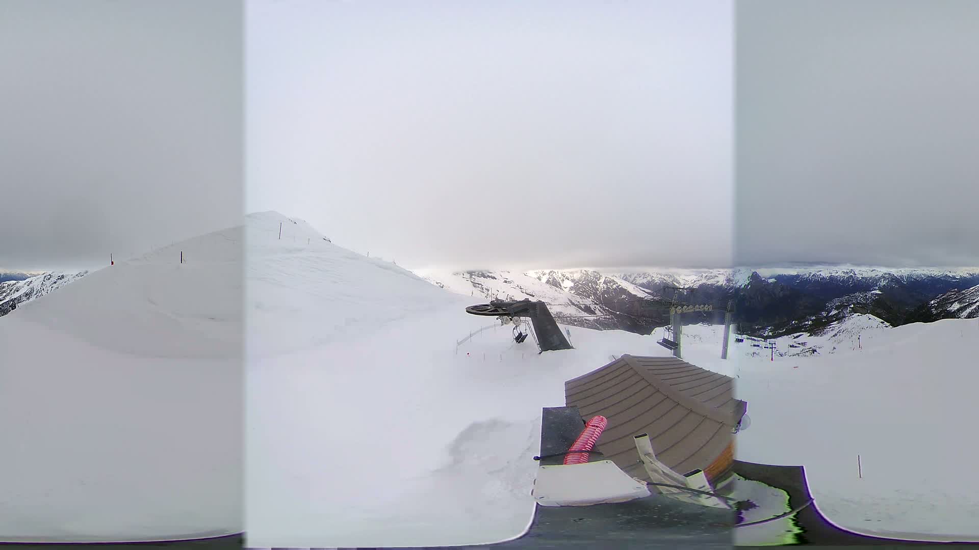 A snow-covered mountaintop view shows a ski lift and a small building under an overcast sky.