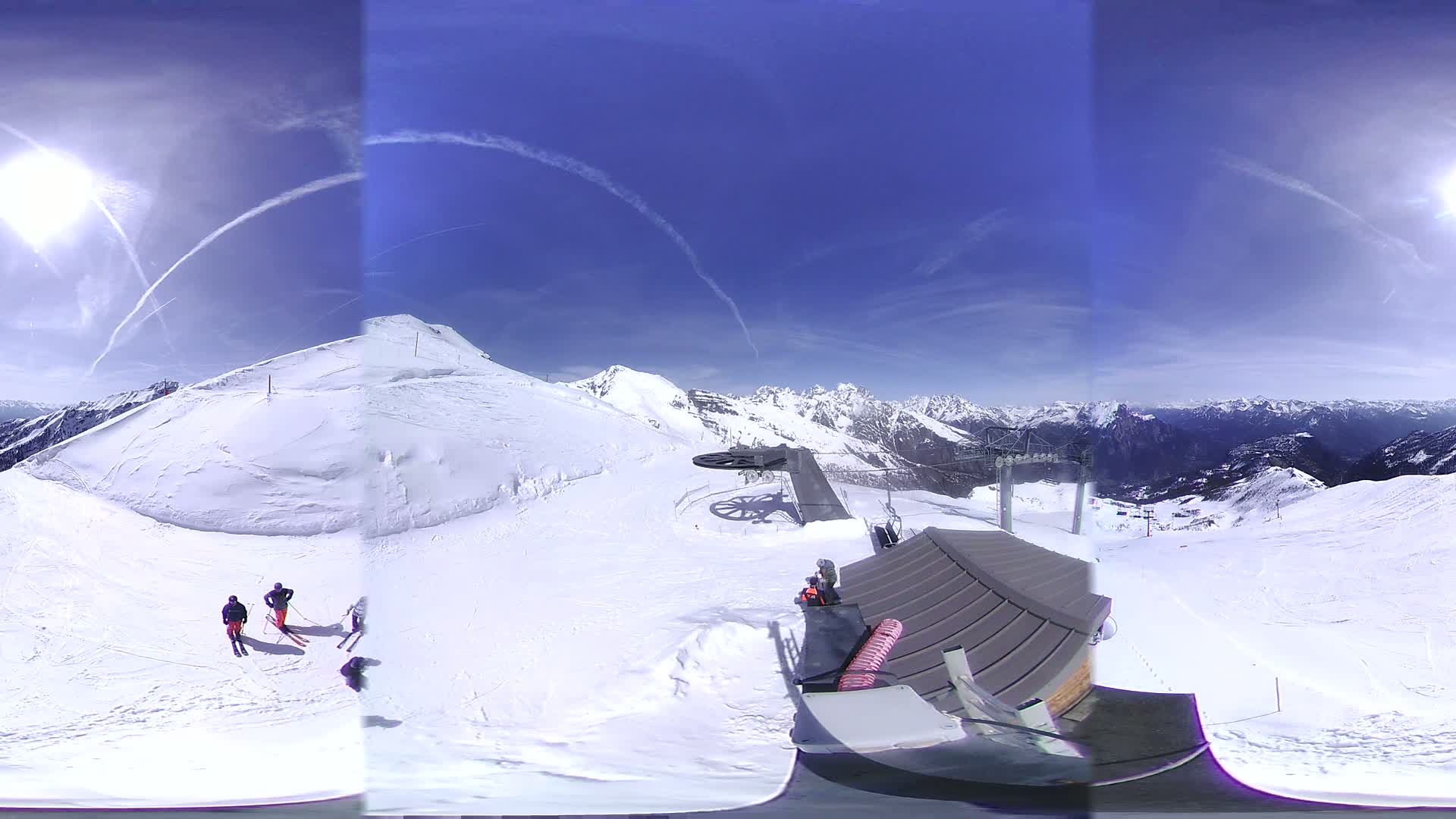 A sunny, snow-covered mountaintop view shows skiers and a ski lift against a backdrop of snow-capped mountains and a mostly clear blue sky.