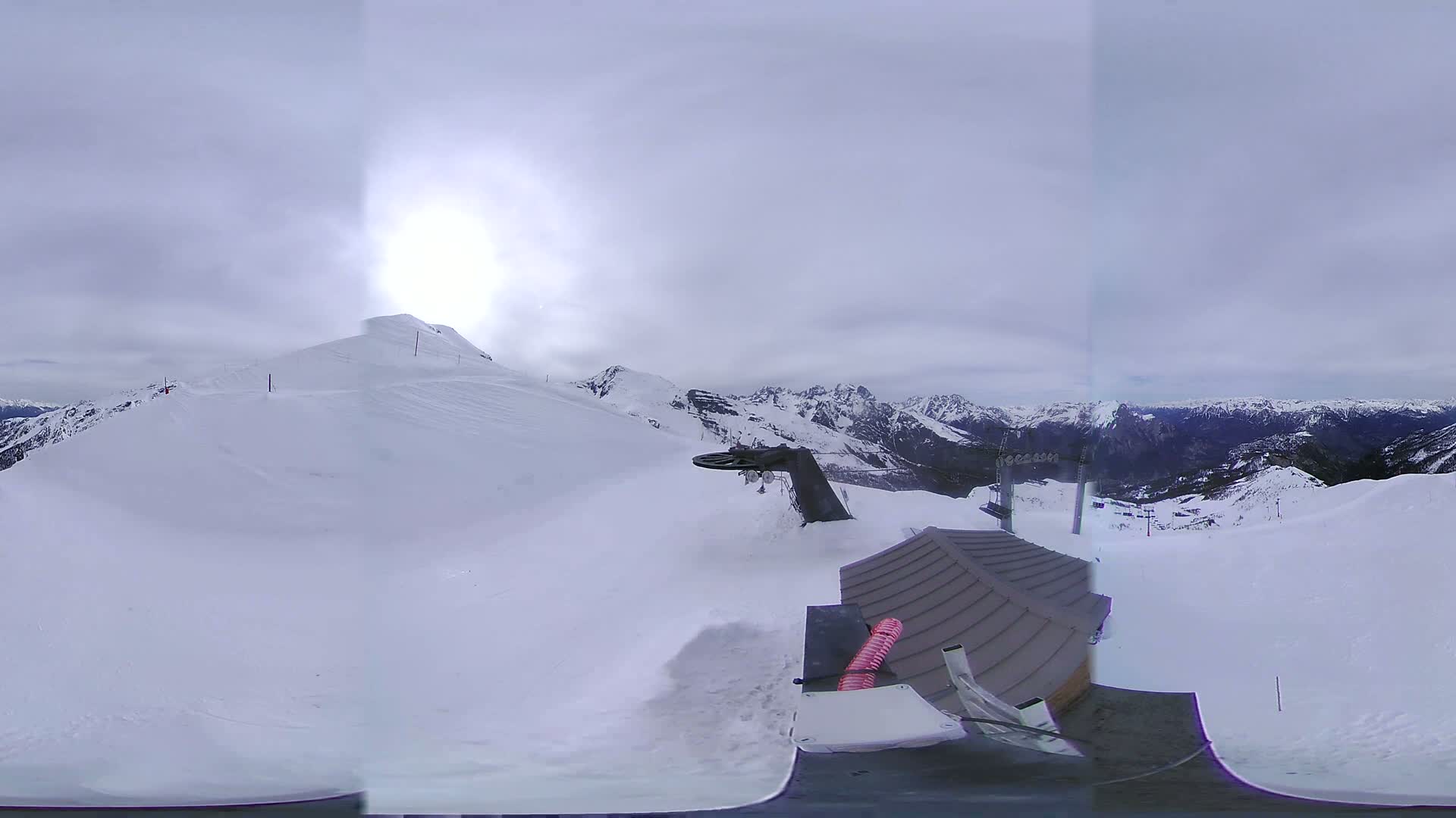A panoramic view from a snowy mountaintop shows a cloudy sky, snow-covered slopes, a ski lift, and a small building.