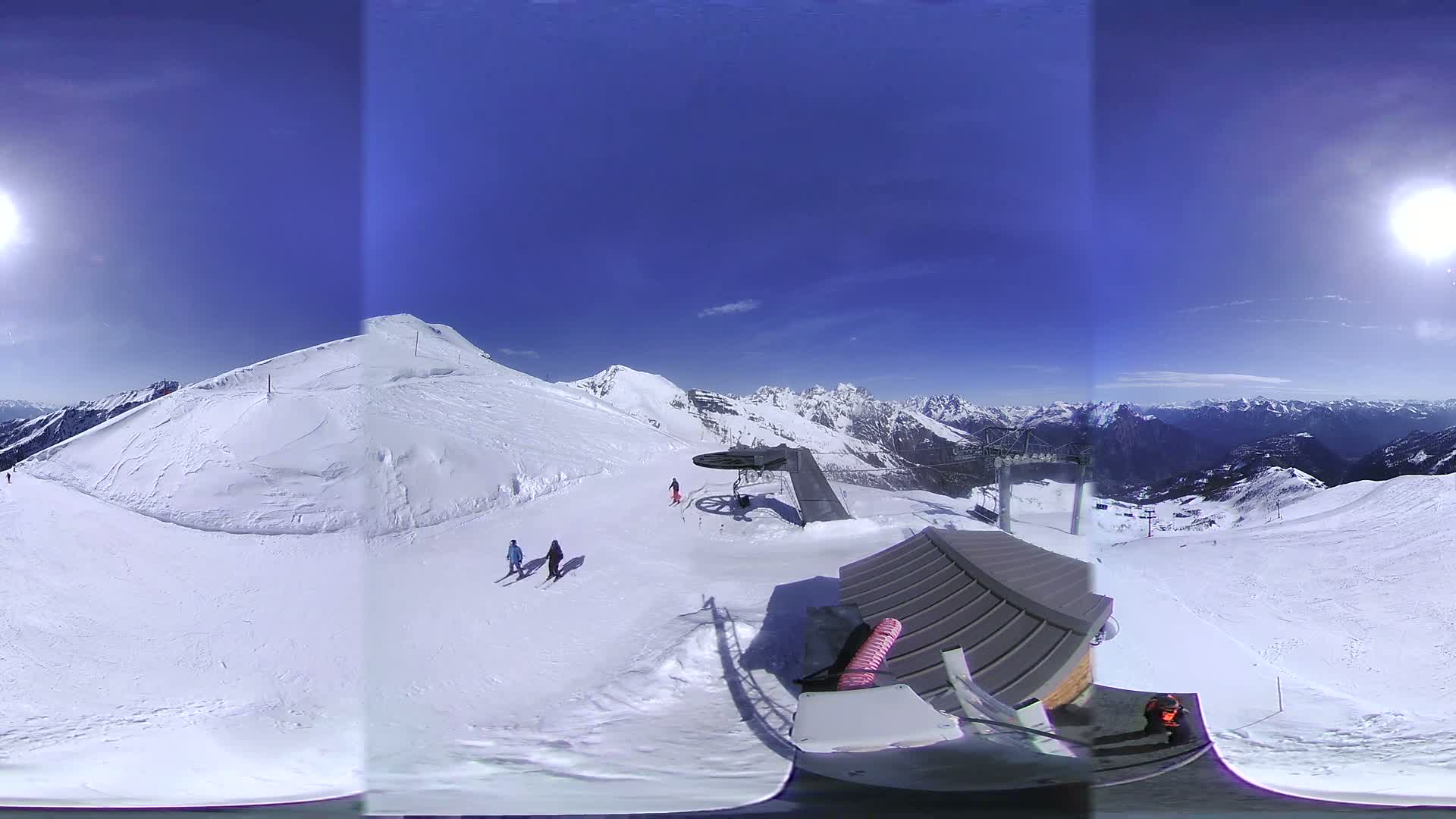A sunny, snow-covered mountaintop ski slope with a ski lift and several skiers visible, overlooking a vast mountain range.