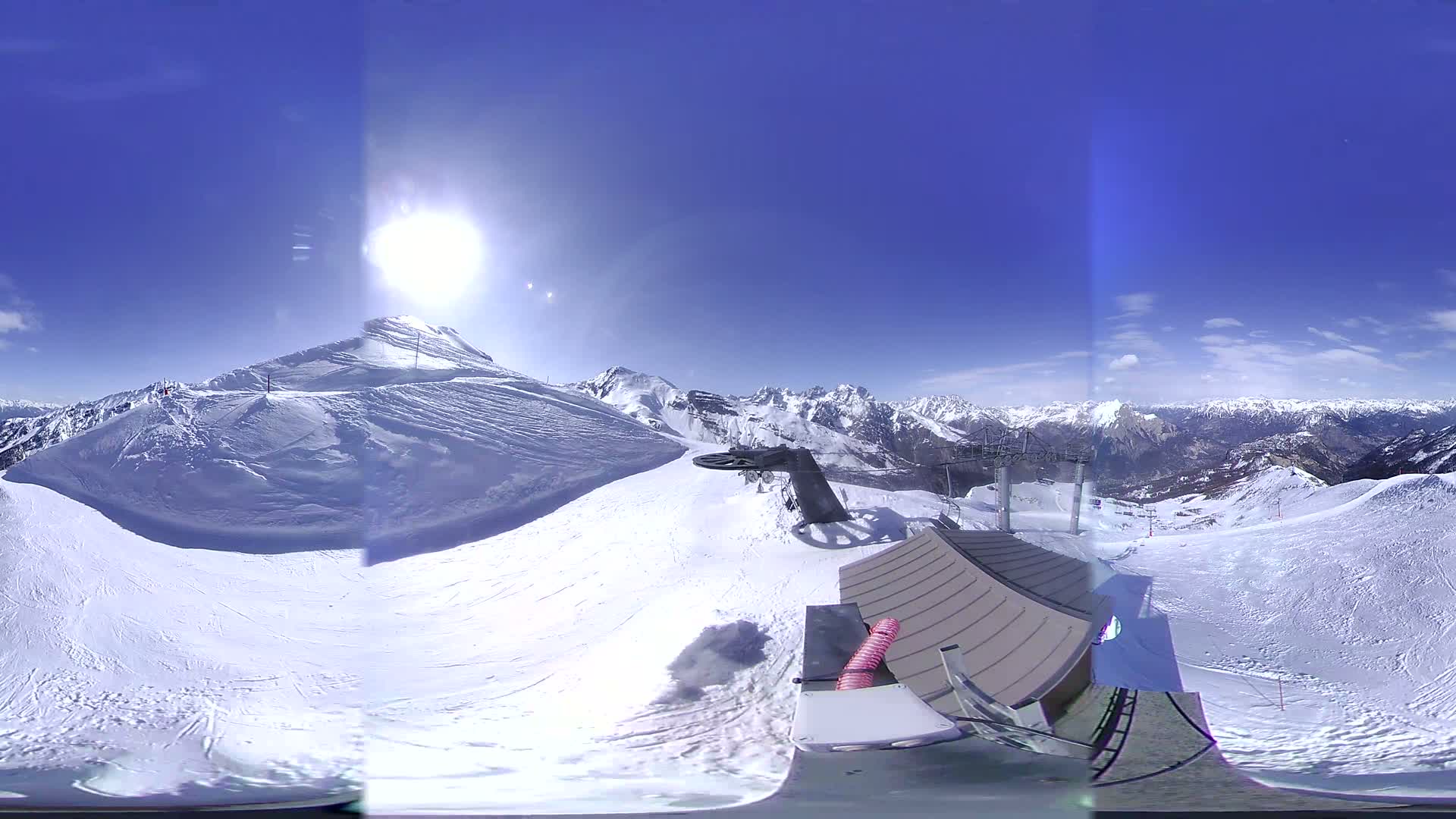 A panoramic view from a snowy mountaintop shows a ski lift, a building, and a bright sunny day with clear blue skies and distant mountain ranges.