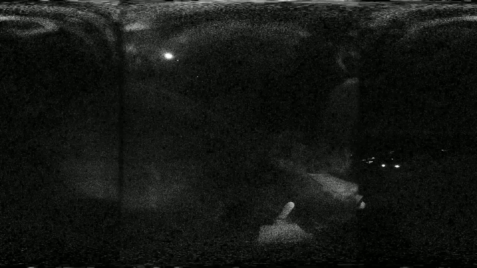 A grainy, nighttime image shows a dimly lit scene with a few faint lights in the distance under a dark sky with a visible moon.