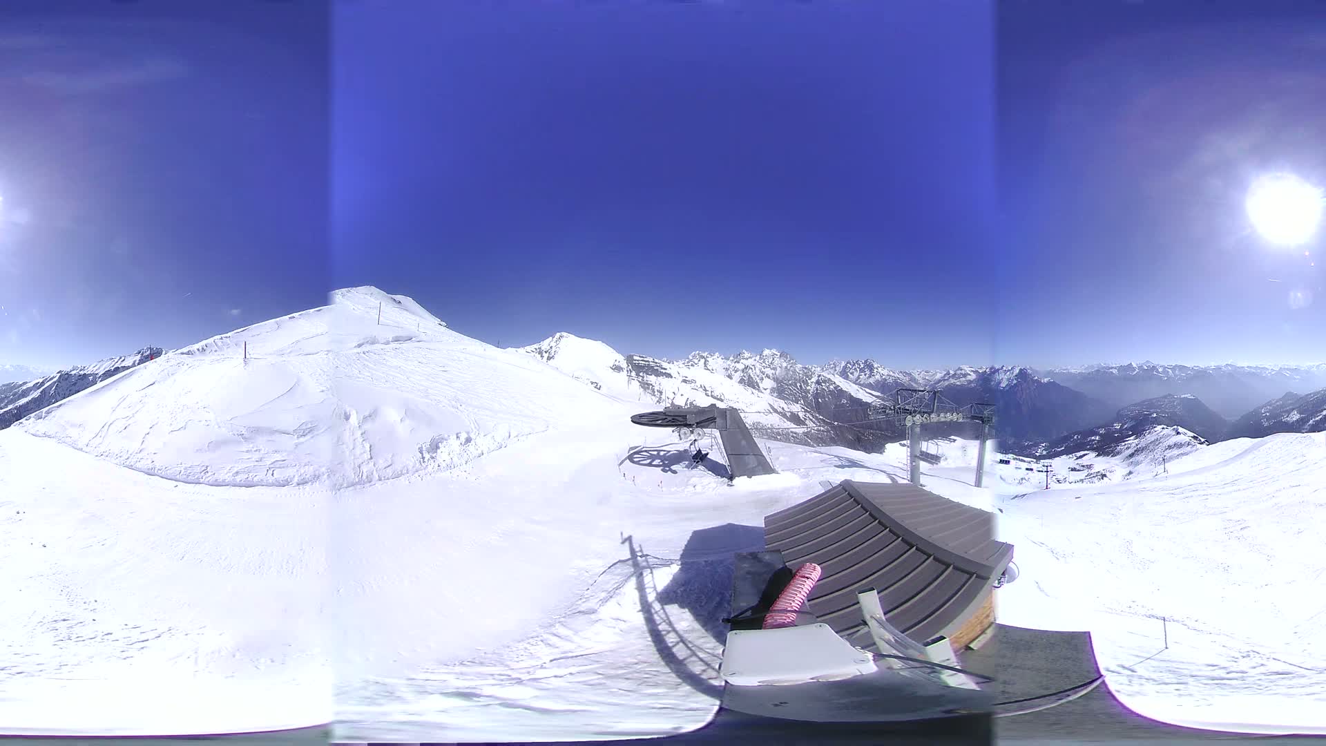 A panoramic view from a snowy mountaintop shows a sunny, clear day with a ski lift and snow-covered slopes and mountains in the background.