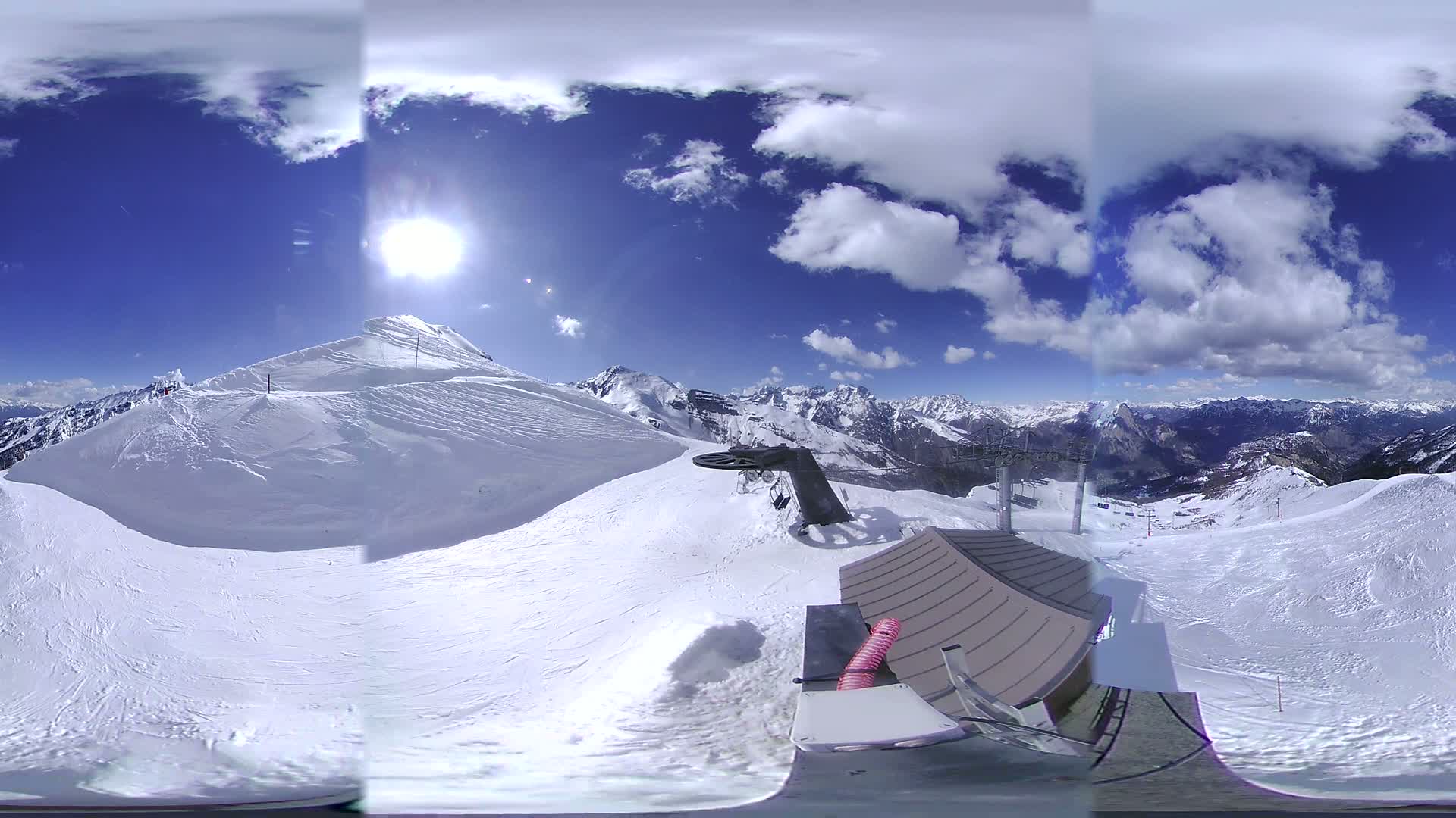 A panoramic view from a snowy mountaintop shows a sunny day with partly cloudy skies, ski slopes, and a ski lift.