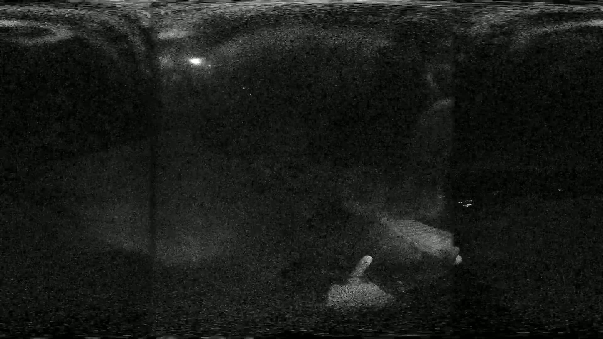 A grainy, low-light nighttime image shows a person standing near a pole with a faint light source in the distance.