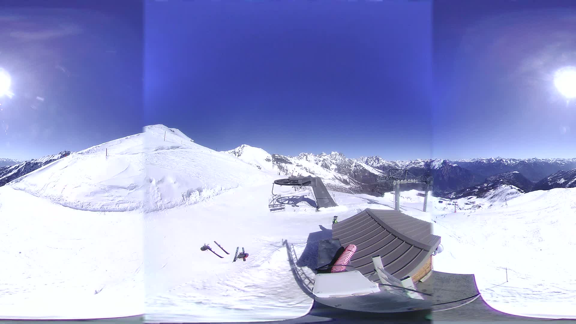 A sunny day on a snow-covered mountain reveals a ski lift, a small building, and a pair of skis discarded on the pristine white slopes, with a mountainous vista in the background.