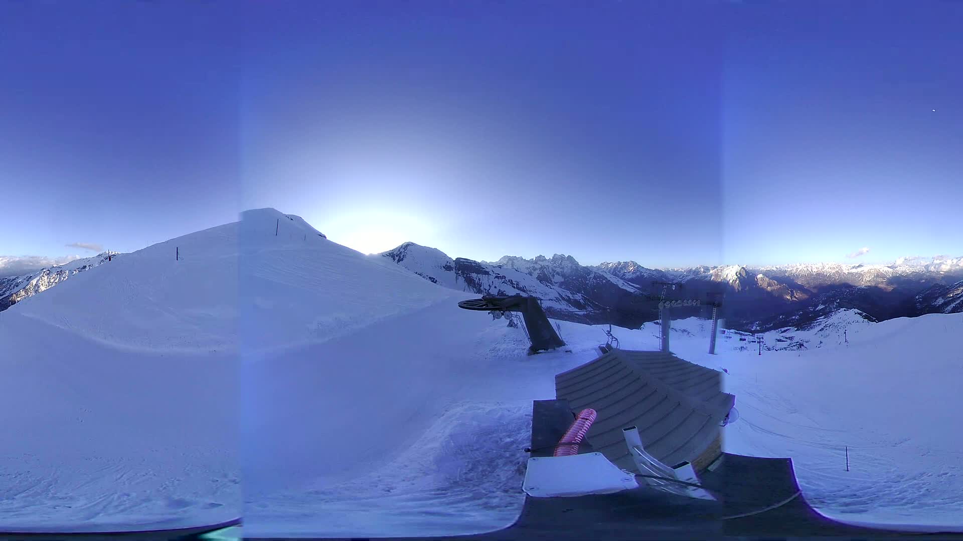 A panoramic view from a snowy mountaintop shows a ski lift, a building, and a vast expanse of snow-covered mountains under a clear blue sky.